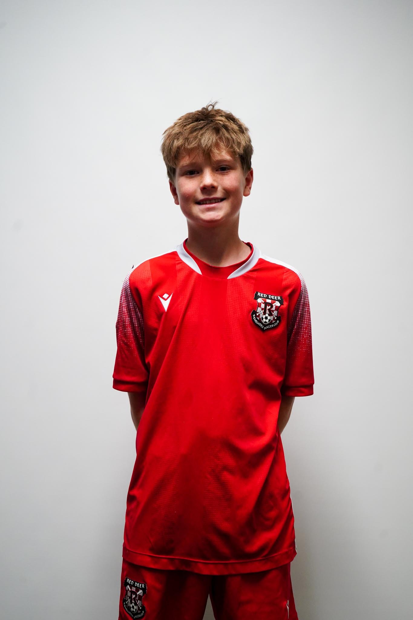 A young boy standing against a plain white background, wearing a red soccer uniform with a crest that reads "Red Deer" and a black and white emblem. He is smiling lightly with his hands behind his back.