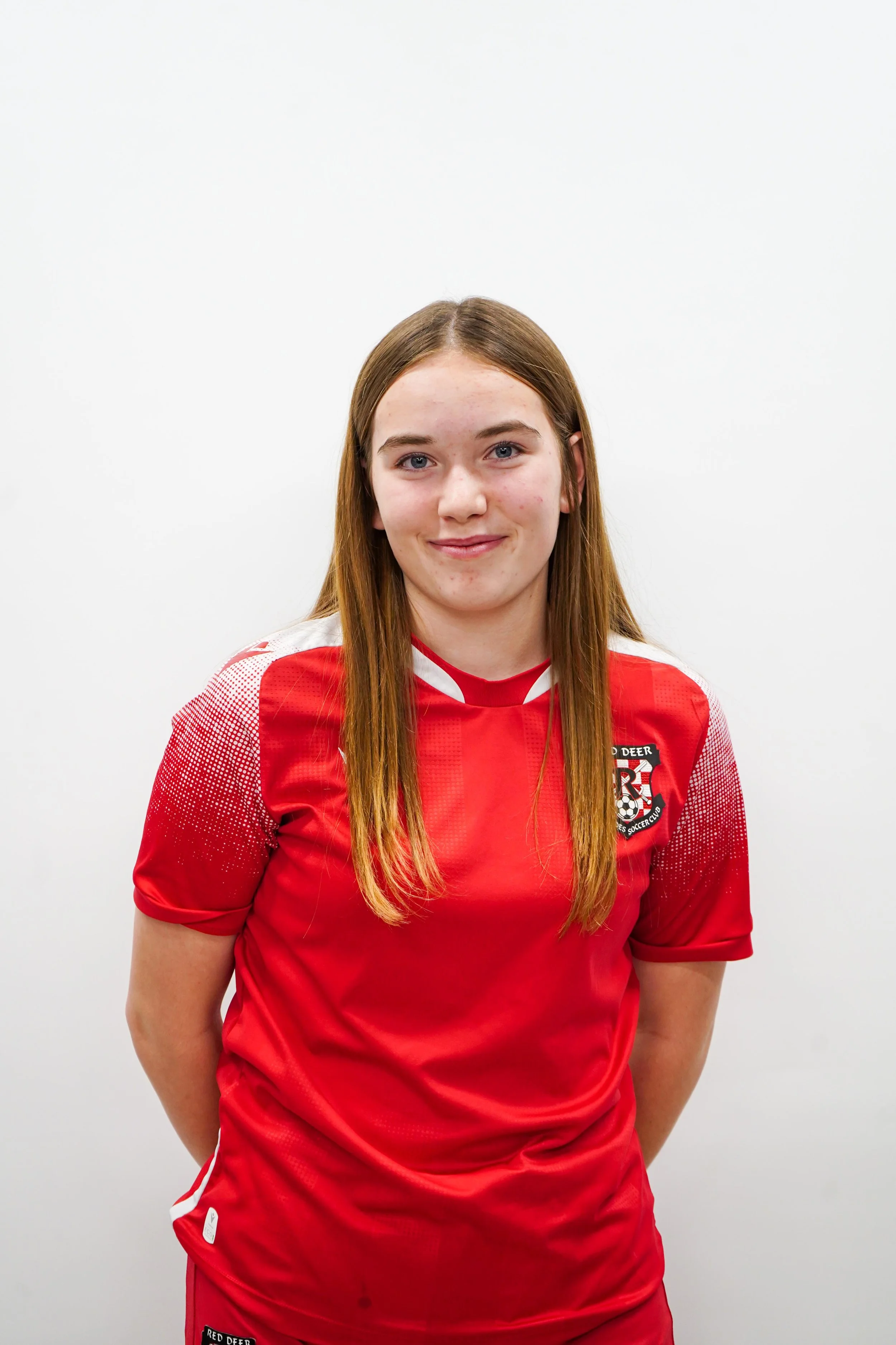A young woman with long red hair wearing a red and white sports jersey standing against a plain white wall.
