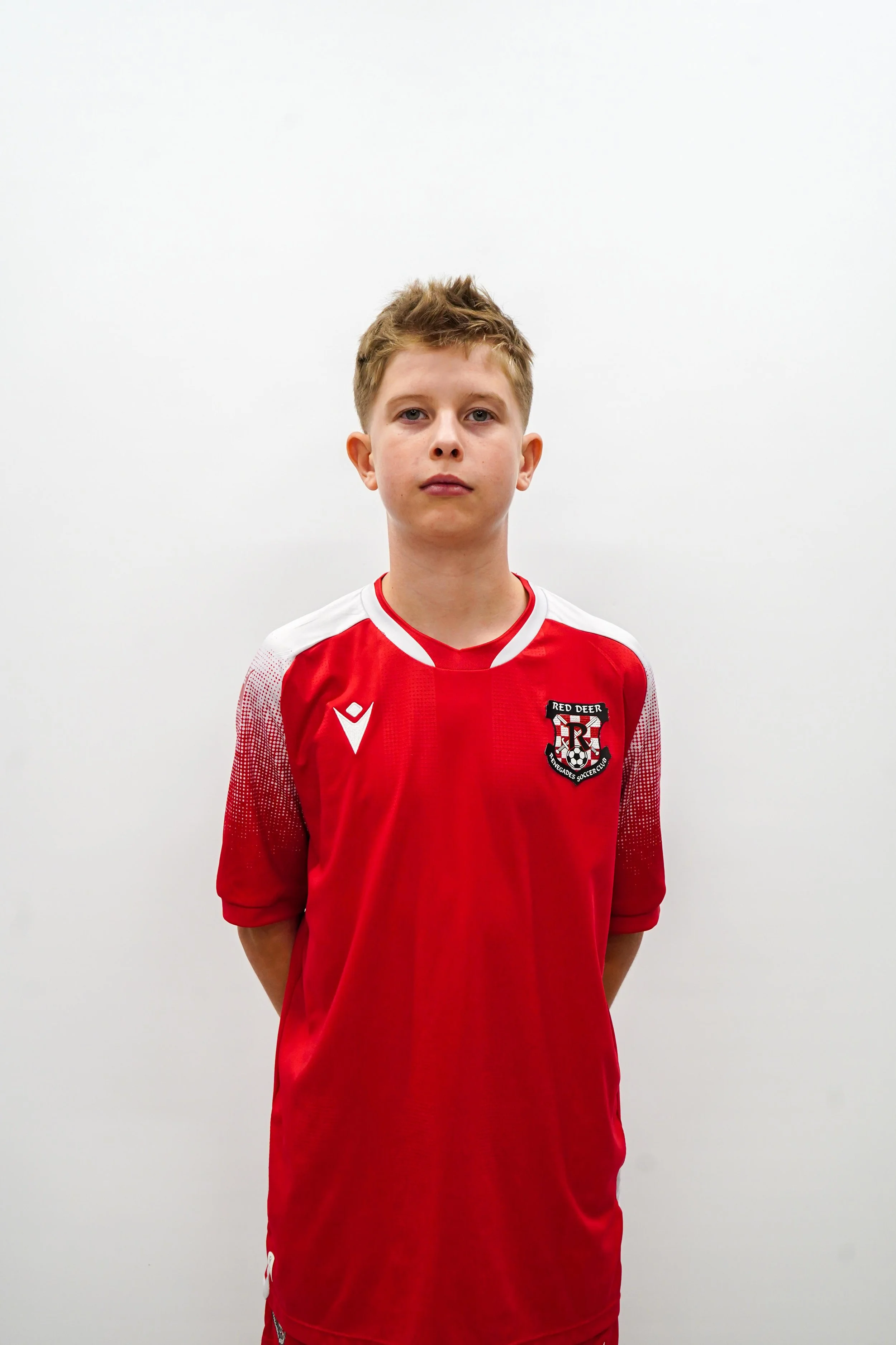 A young boy wearing a red soccer jersey with white accents and a crest on the chest, standing against a plain white background.