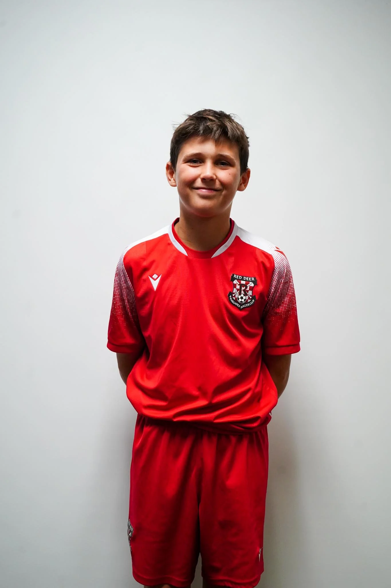 A young boy wearing a red soccer uniform standing in front of a plain white wall.