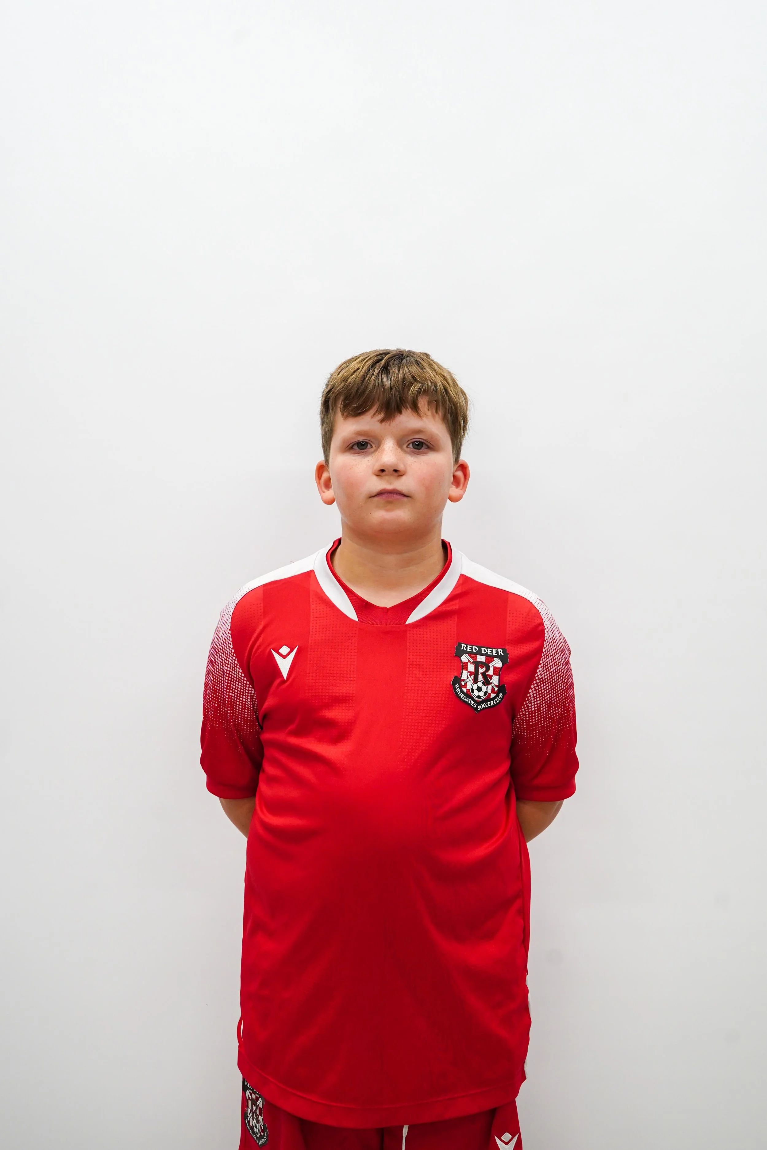 A young boy in a red soccer jersey with a crest on the chest stands against a plain white background.