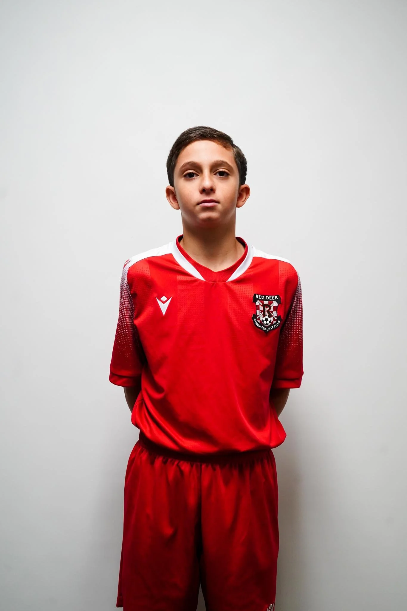 Young boy wearing a red soccer uniform standing against a plain white wall.