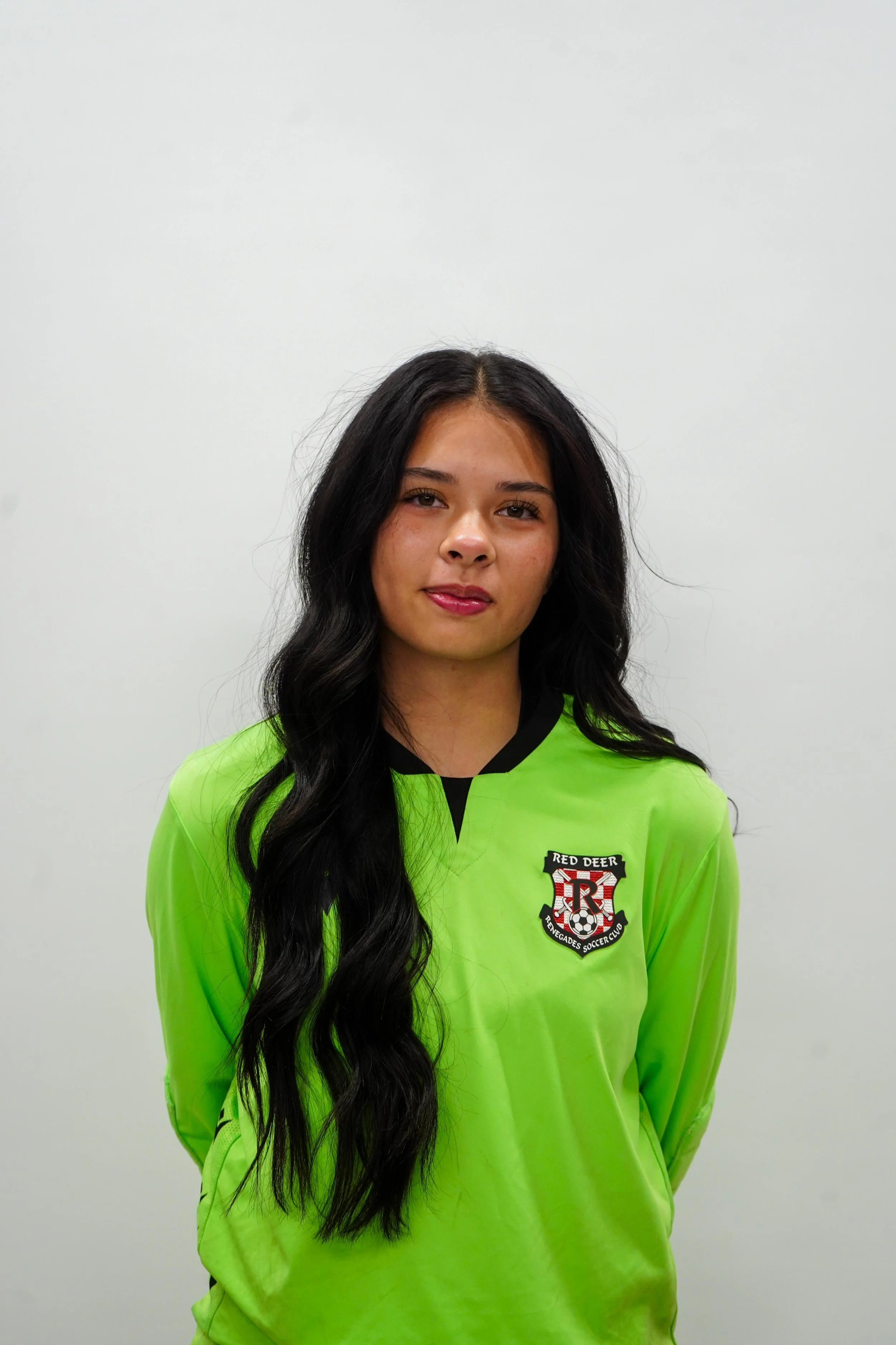 A young woman with long black hair in a yellow-green soccer jersey with the Red Deer Uncaged Soccer Club logo, standing against a plain white background.