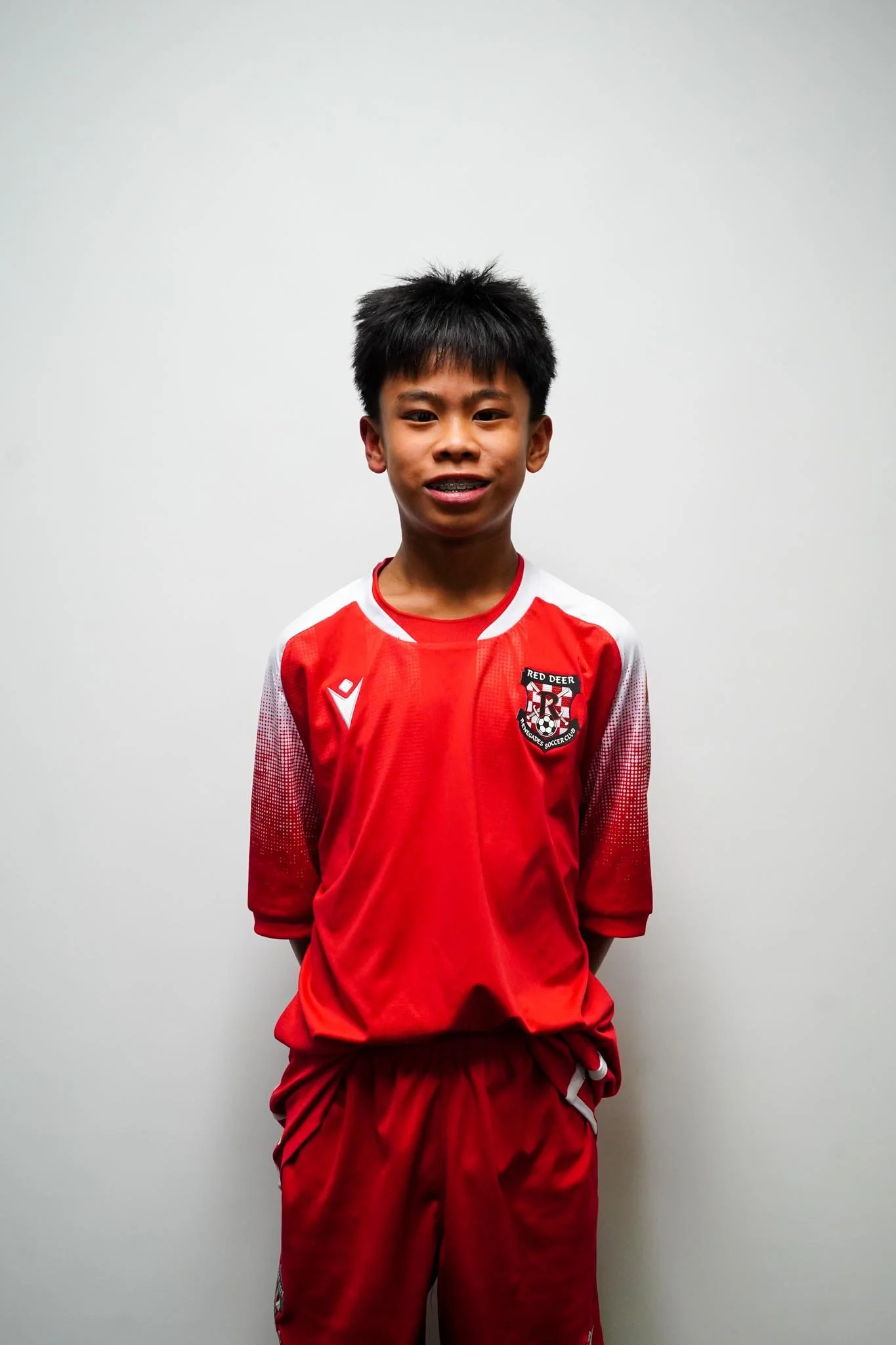 Young boy wearing a red soccer uniform with a team crest on the chest, standing against a plain white wall.