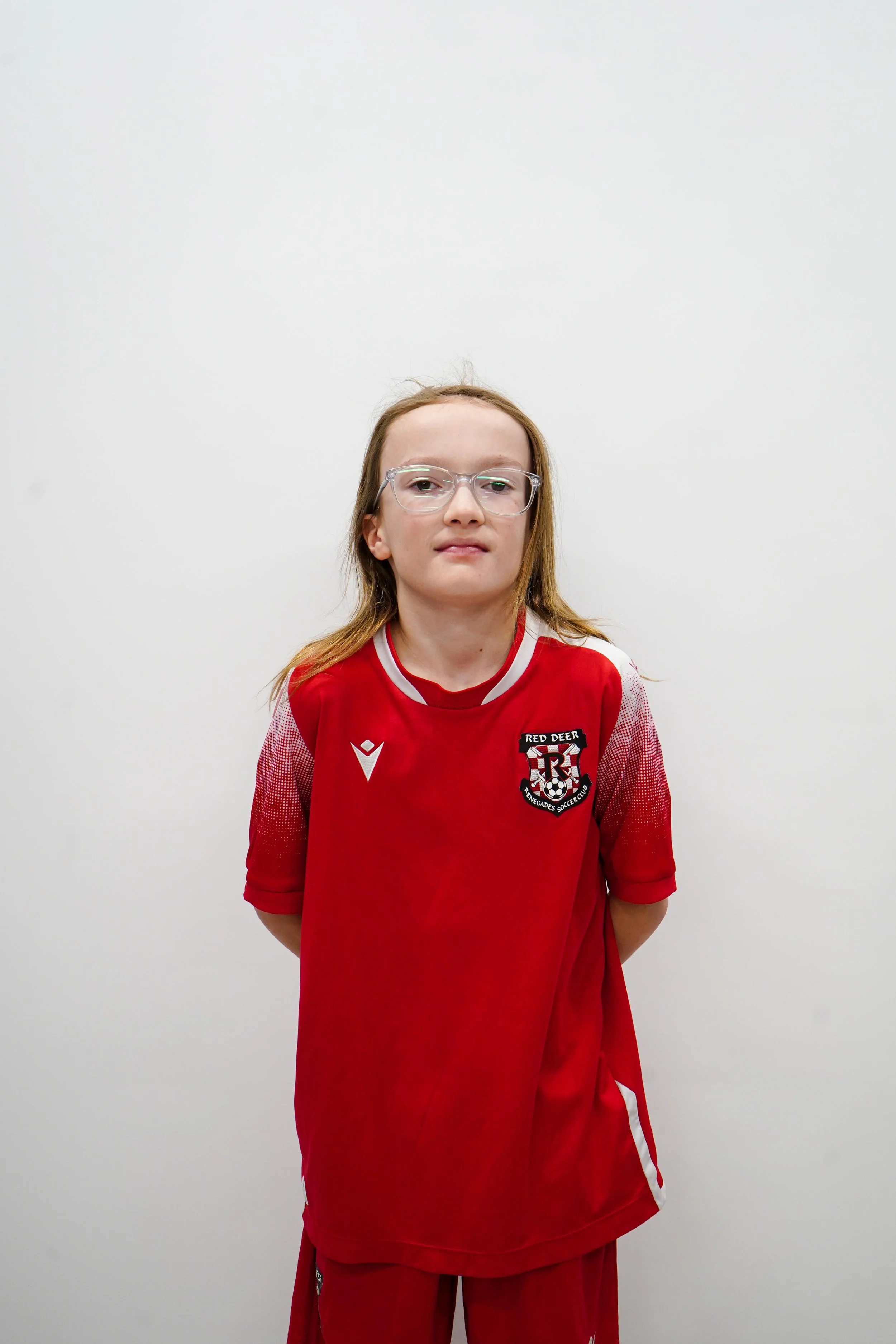 Young girl in a red sports jersey with glasses standing against a plain white wall.