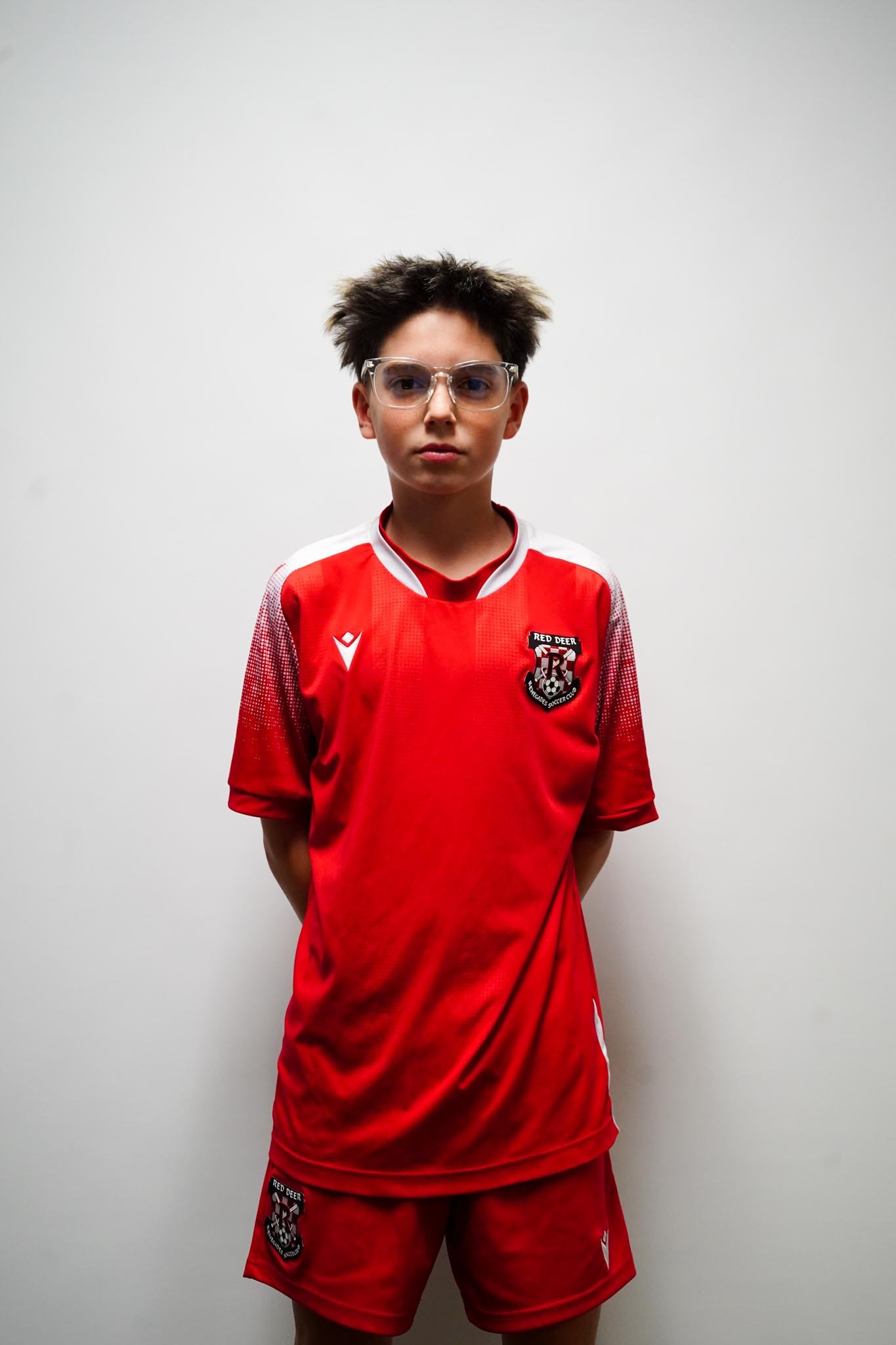A young boy wearing a red soccer uniform with glasses, standing against a plain white background.