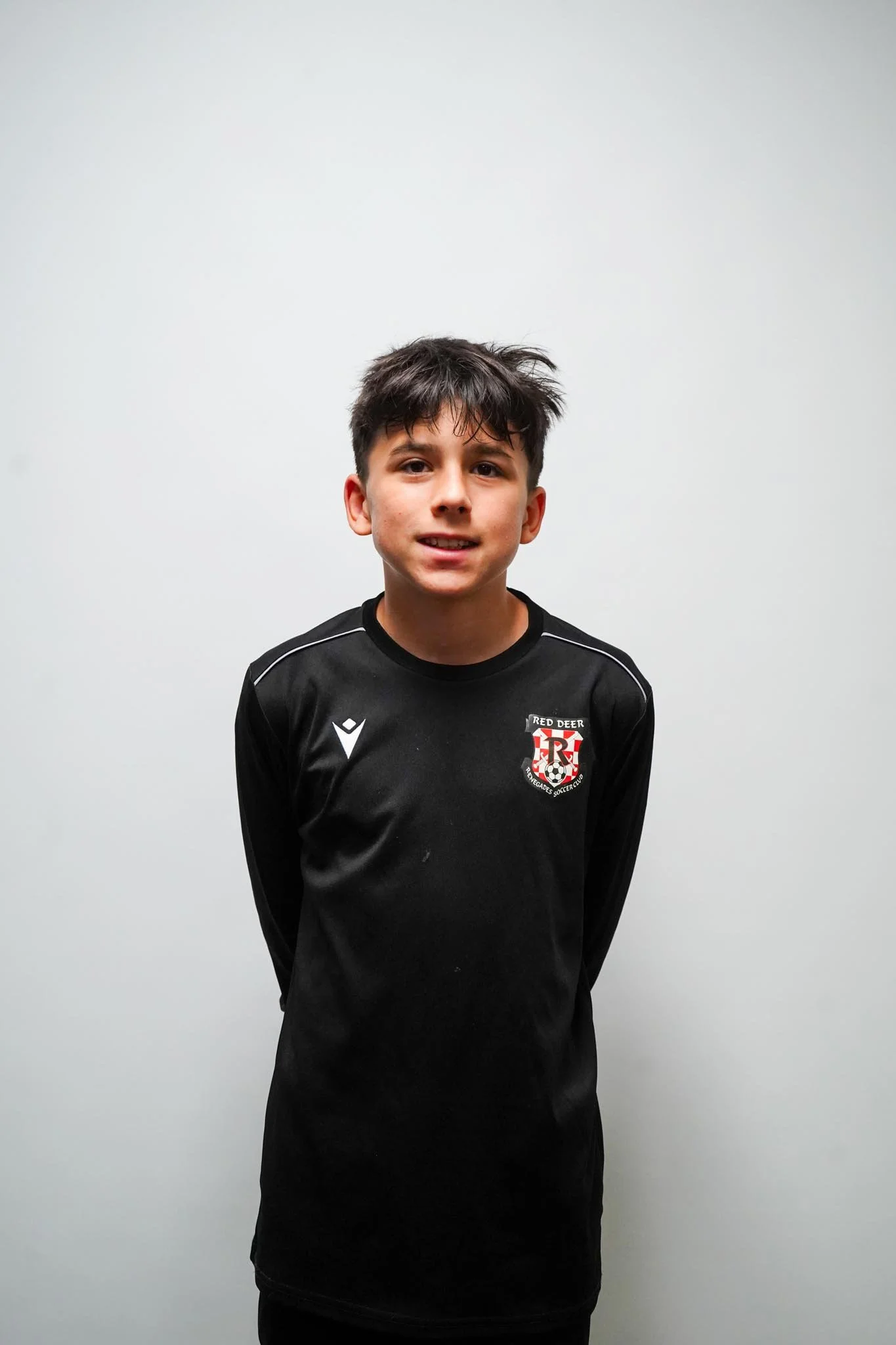 Young boy with dark hair wearing a black soccer jersey standing against a plain white wall.