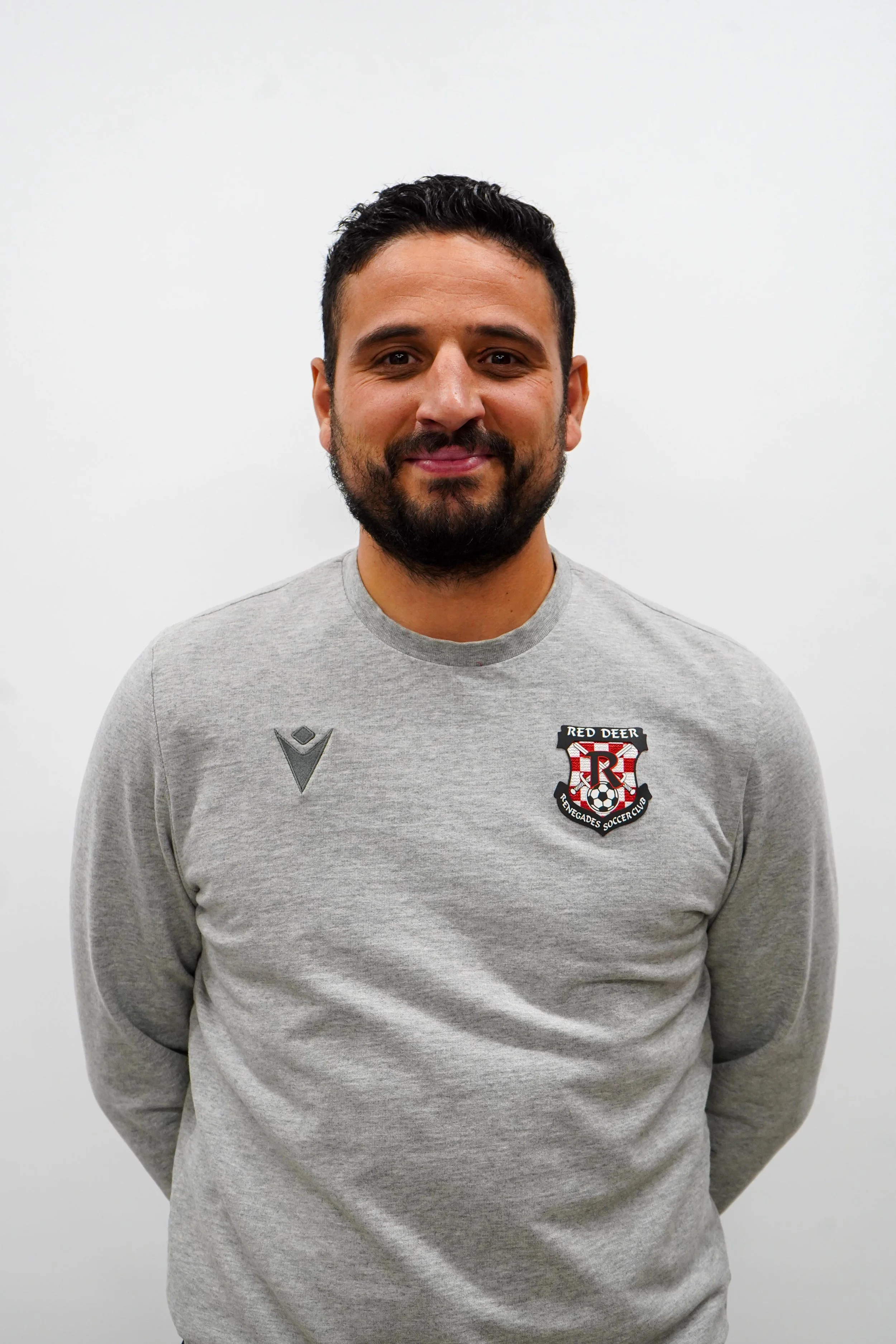 A man with dark hair, beard, and mustache standing against a plain white wall, smiling, wearing a gray sweatshirt with a 'Red Deer Renegades Soccer Club' logo on the left chest.
