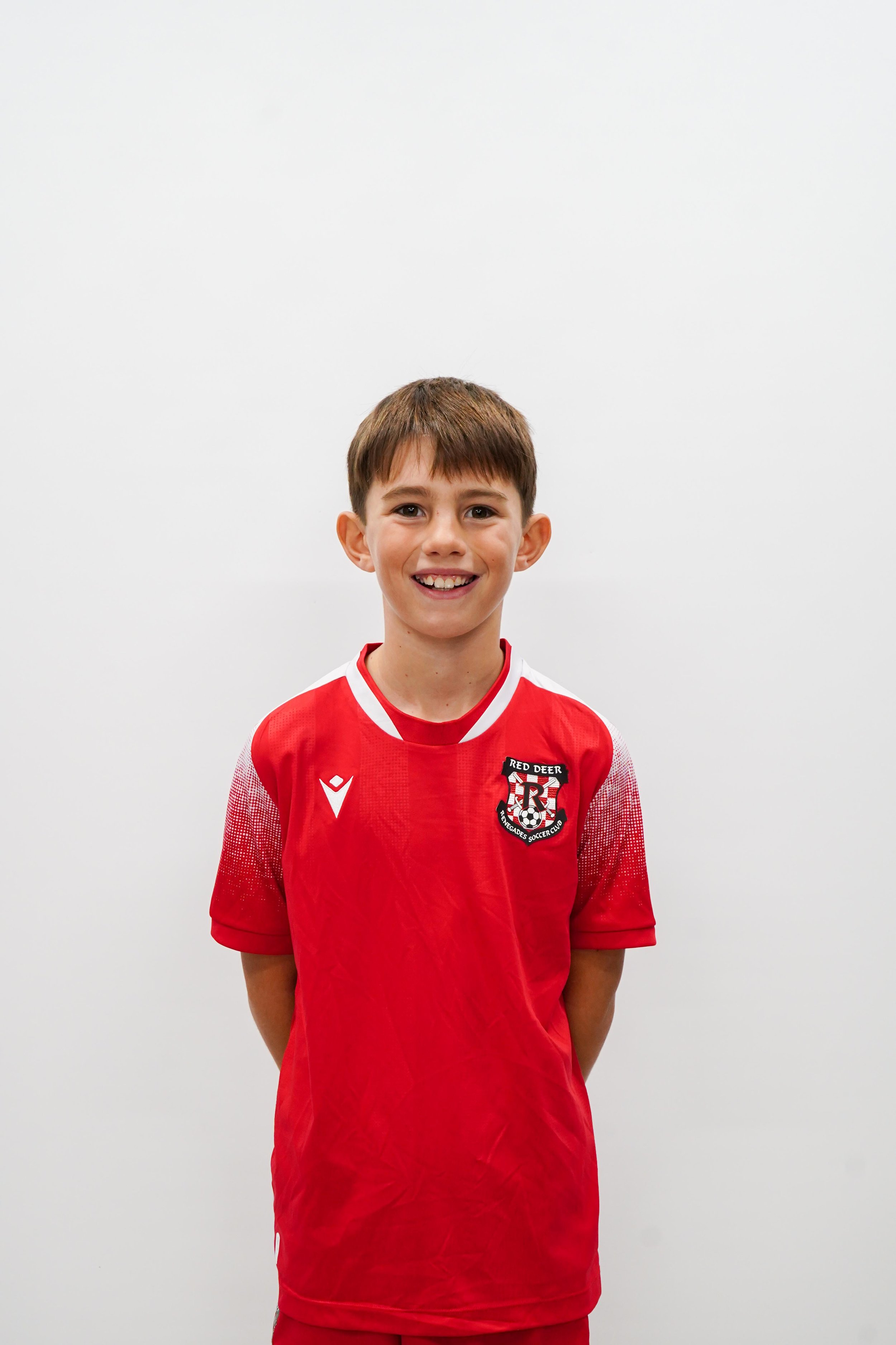 Young boy wearing a red soccer jersey with a logo on it, smiling and standing against a white background.