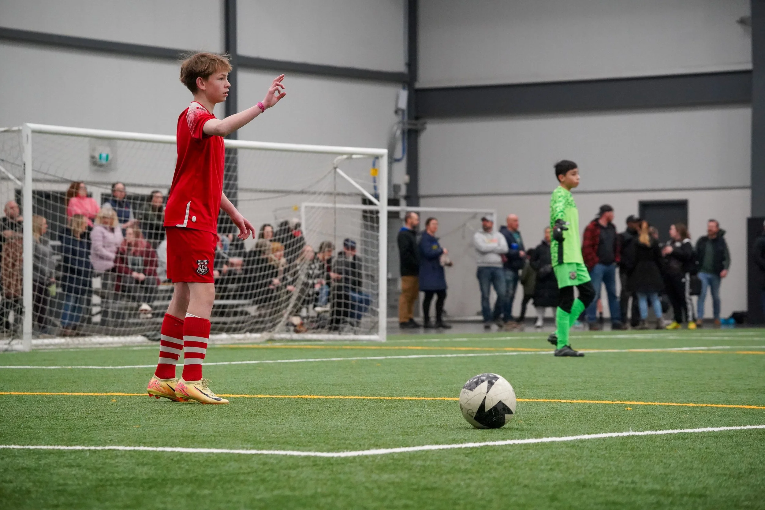 Red Deer Renegades soccer players on an indoor field, one in a red uniform preparing to take a free kick, and the goalkeeper in a green uniform standing near the goal, with spectators watching in the background.