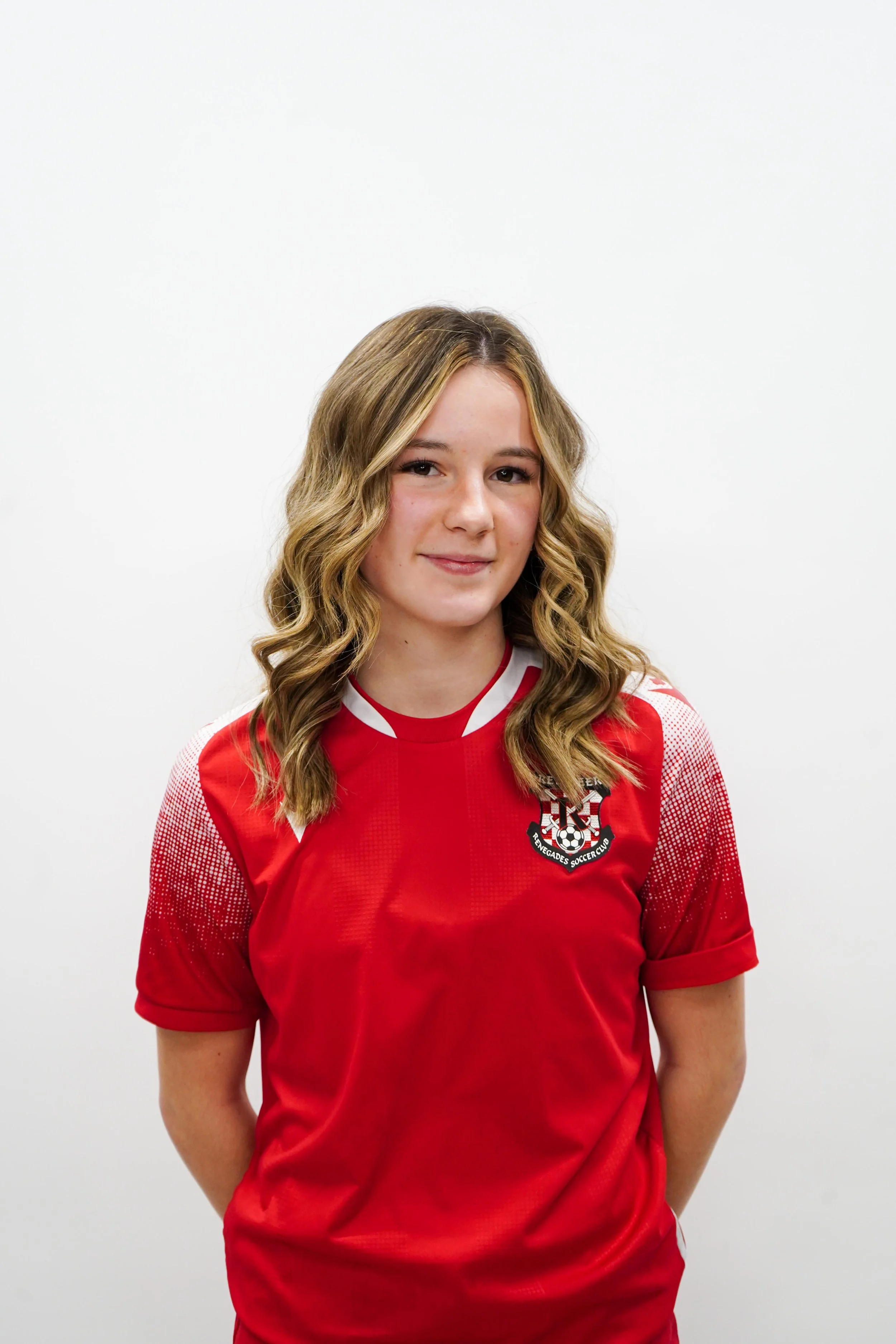 Young woman with wavy blonde hair wearing a red sports jersey with a soccer team logo, standing against a plain white background.
