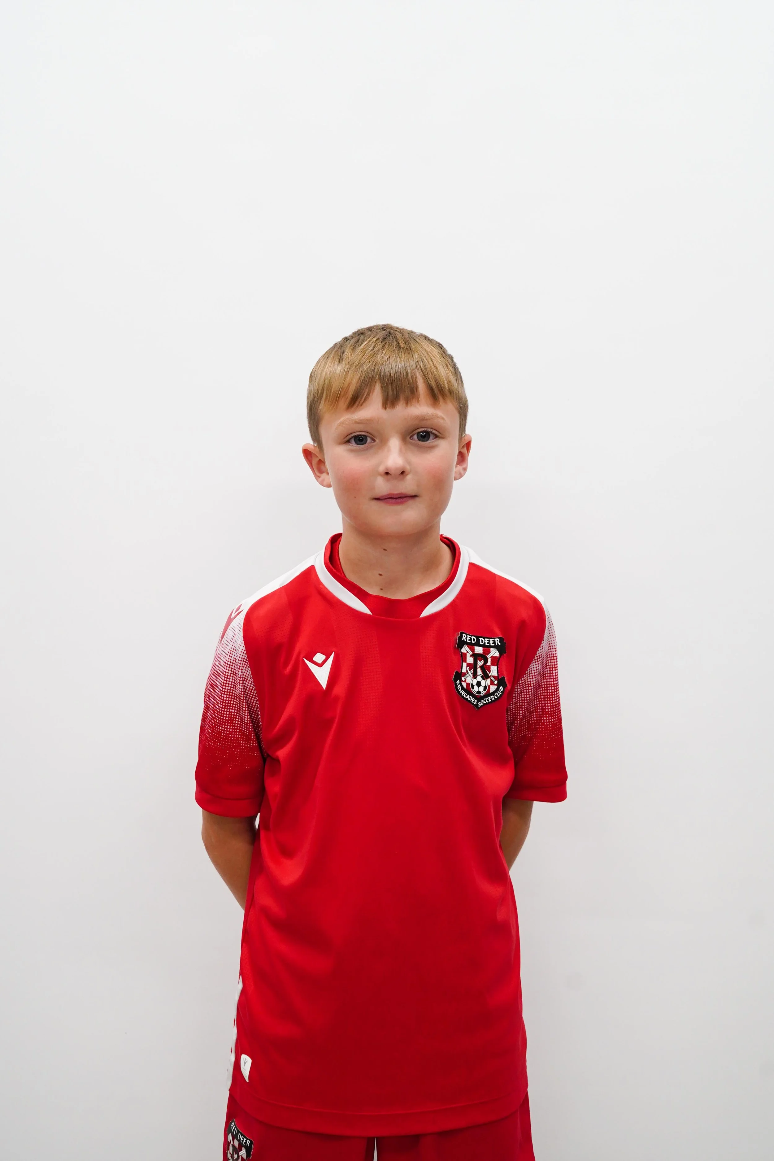 Young boy in a red soccer jersey standing against a plain white wall.