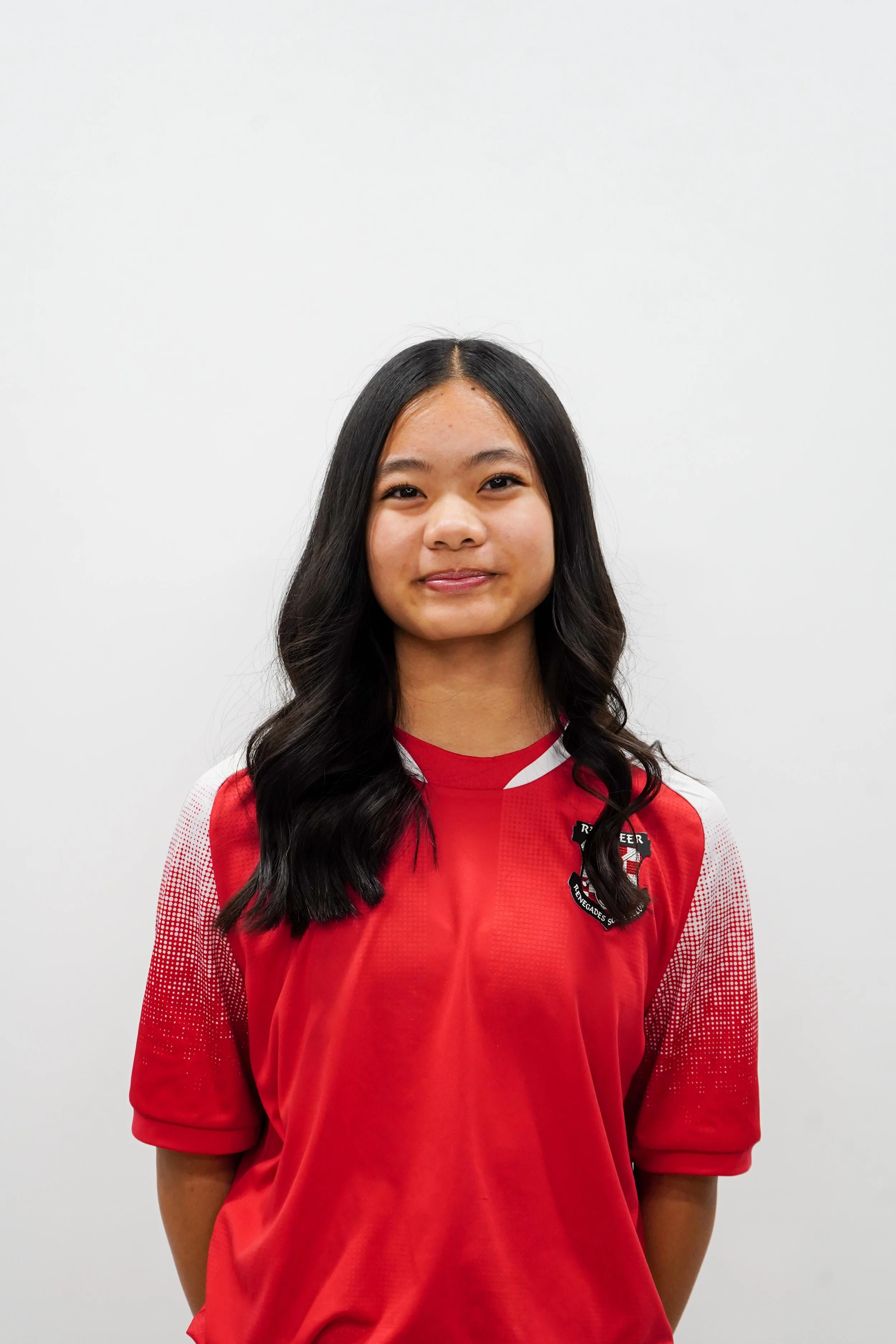 A girl with long black wavy hair wearing a red sports jersey with a black and white logo on the chest standing against a plain white background.