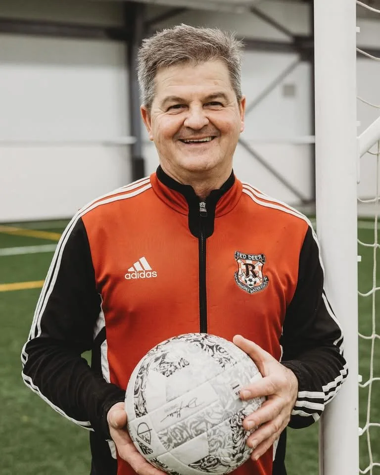 A man smiling while holding a soccer ball on a soccer field.