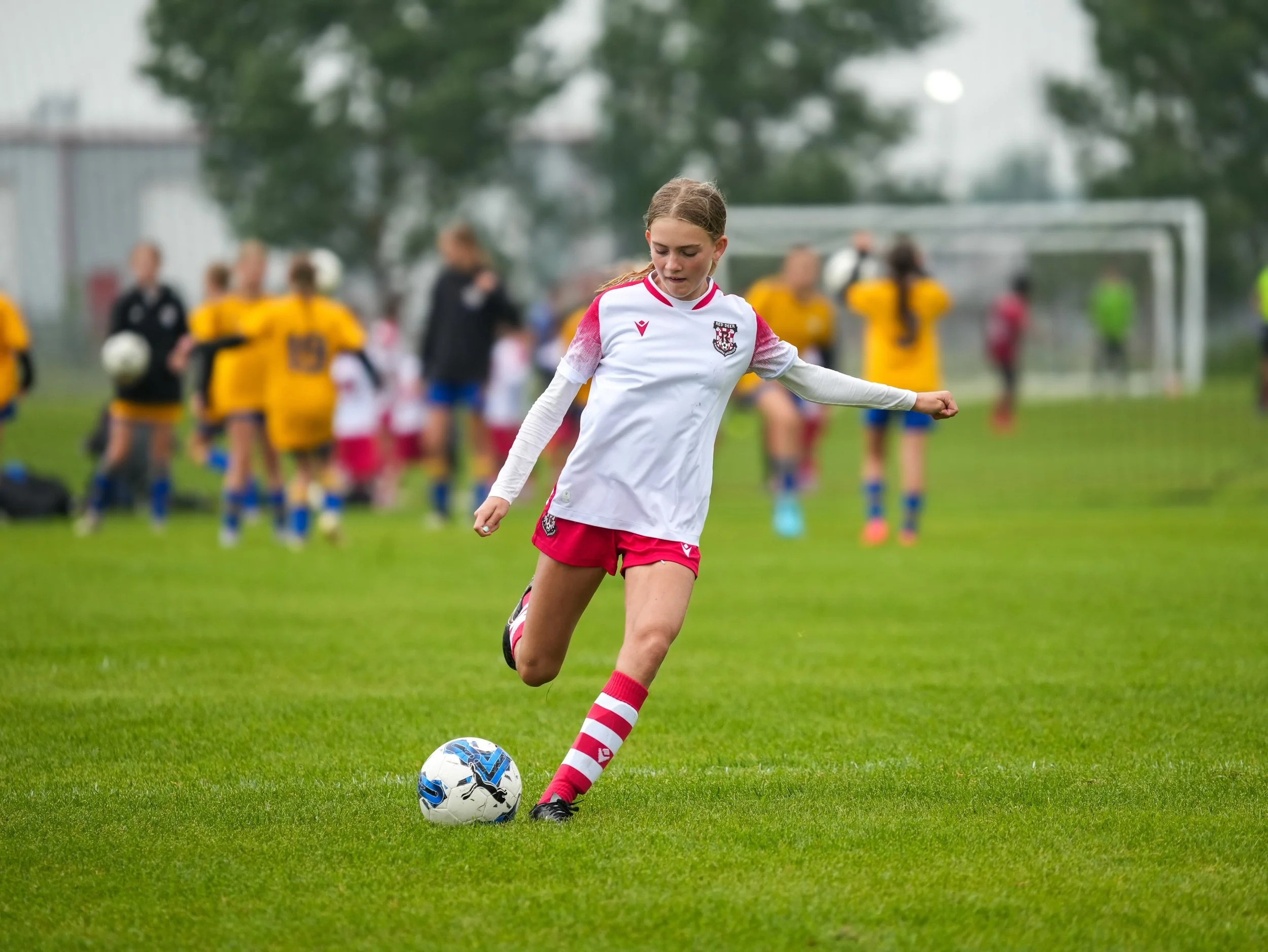 A young girl in a white and red soccer uniform kicks a soccer ball on a grassy field during a match, with other players and a football goal in the background.