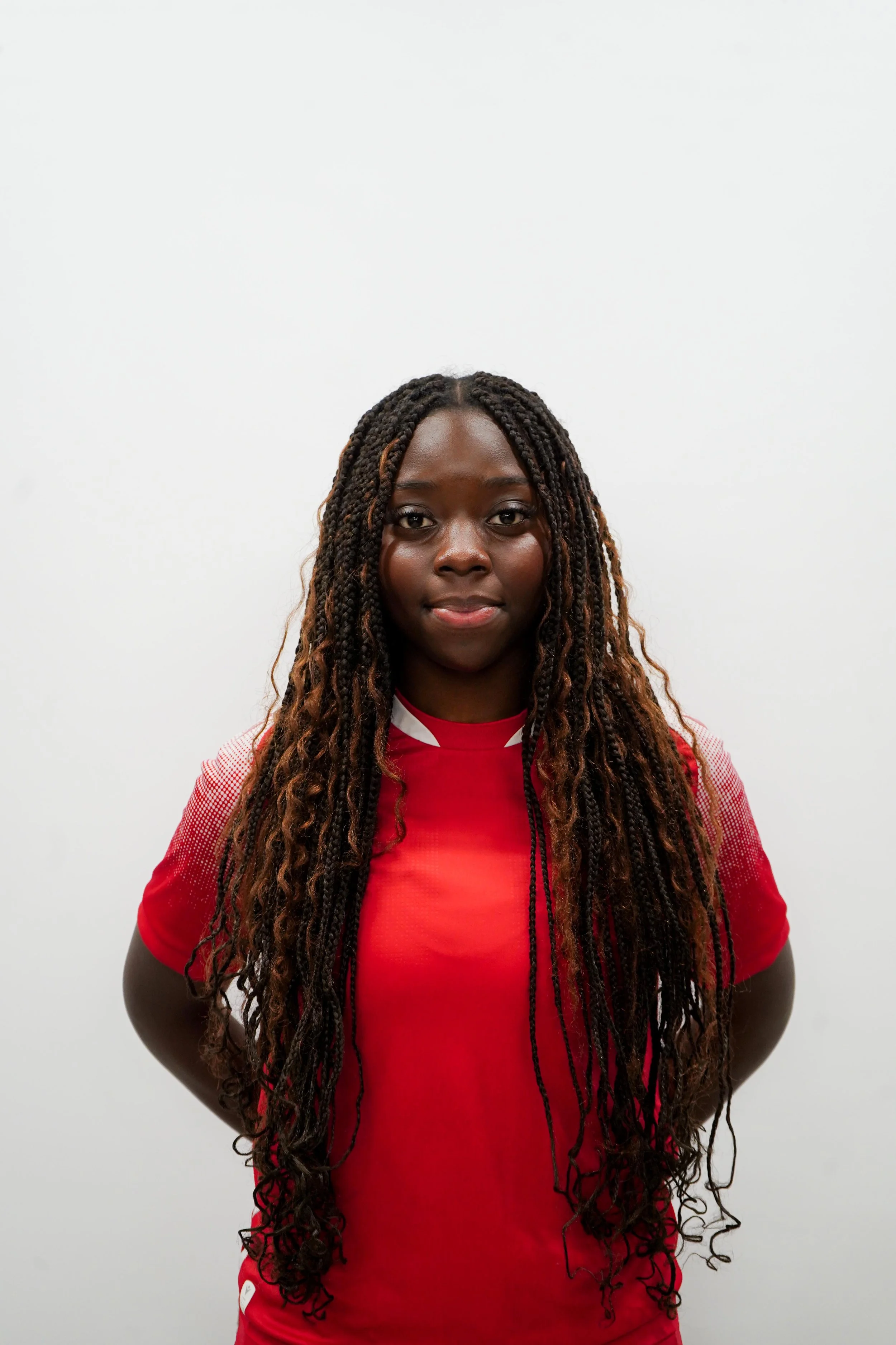 A young woman with long, twisted braids wearing a red athletic shirt, standing against a plain white background.