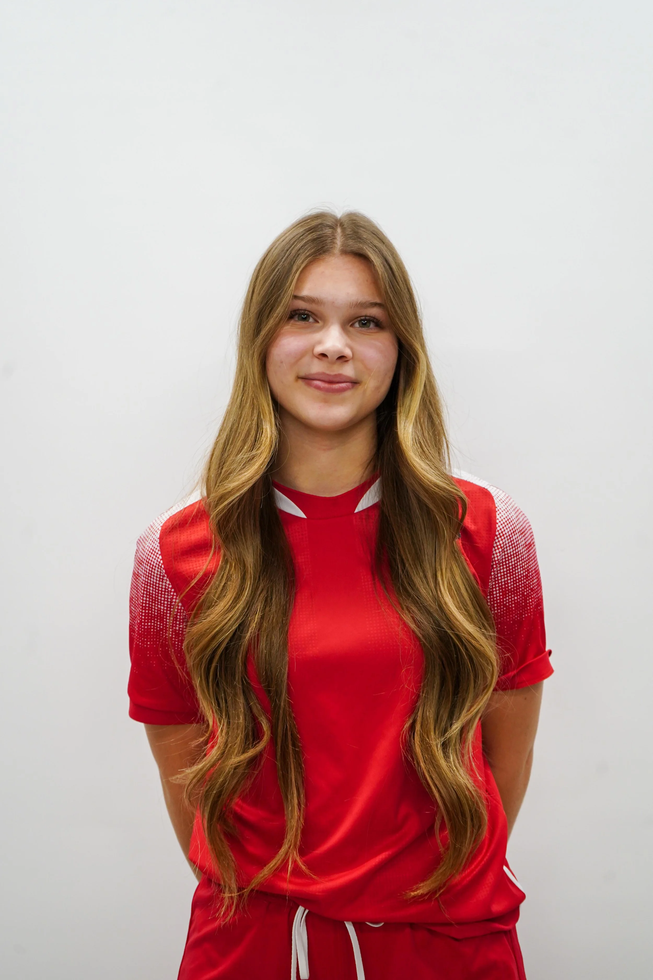A young woman with long wavy blonde hair, wearing a red sports jersey, standing against a plain white wall.