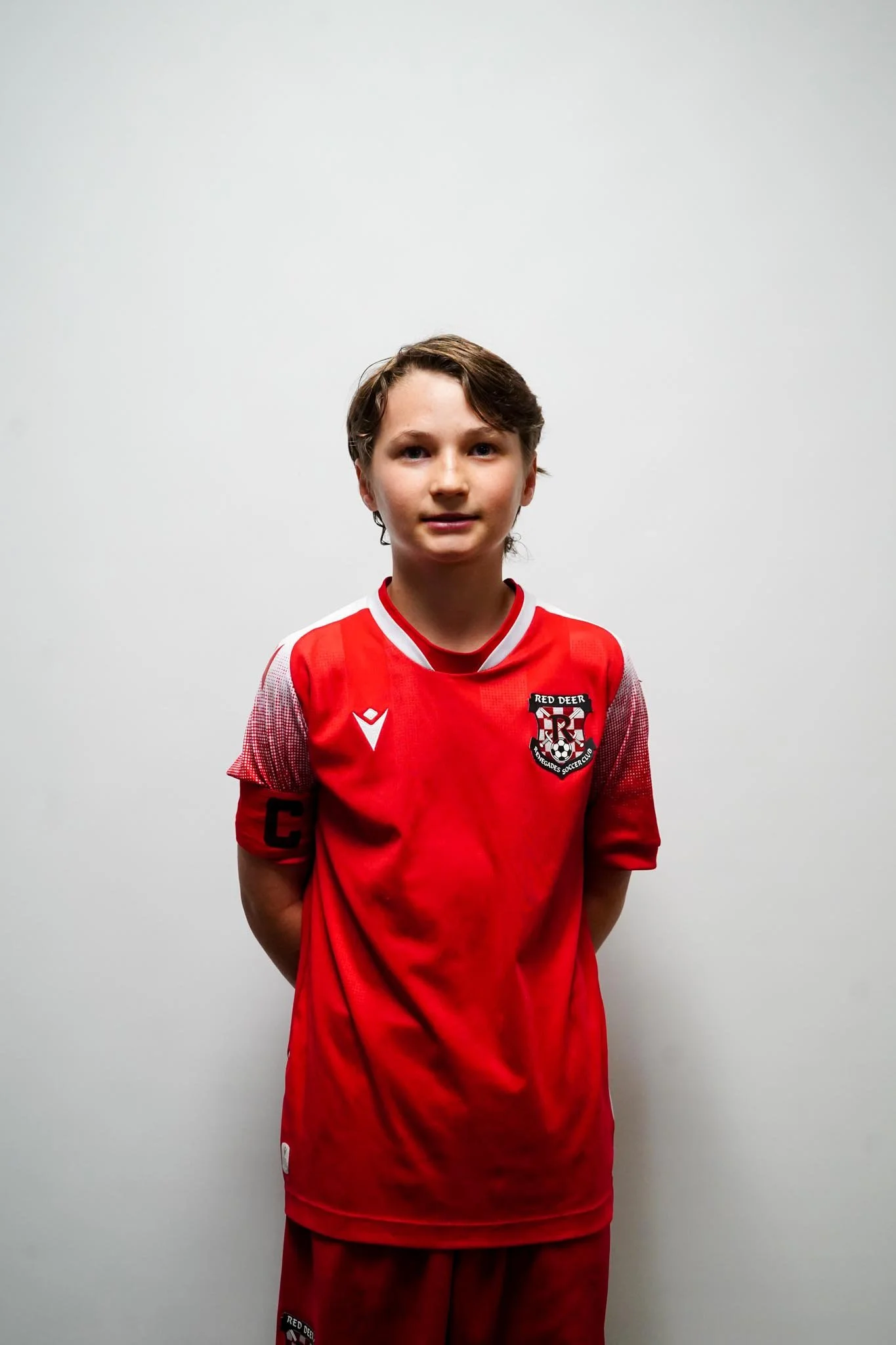 A young boy wearing a red soccer jersey with a 'Red Deer' logo, standing against a plain white wall.