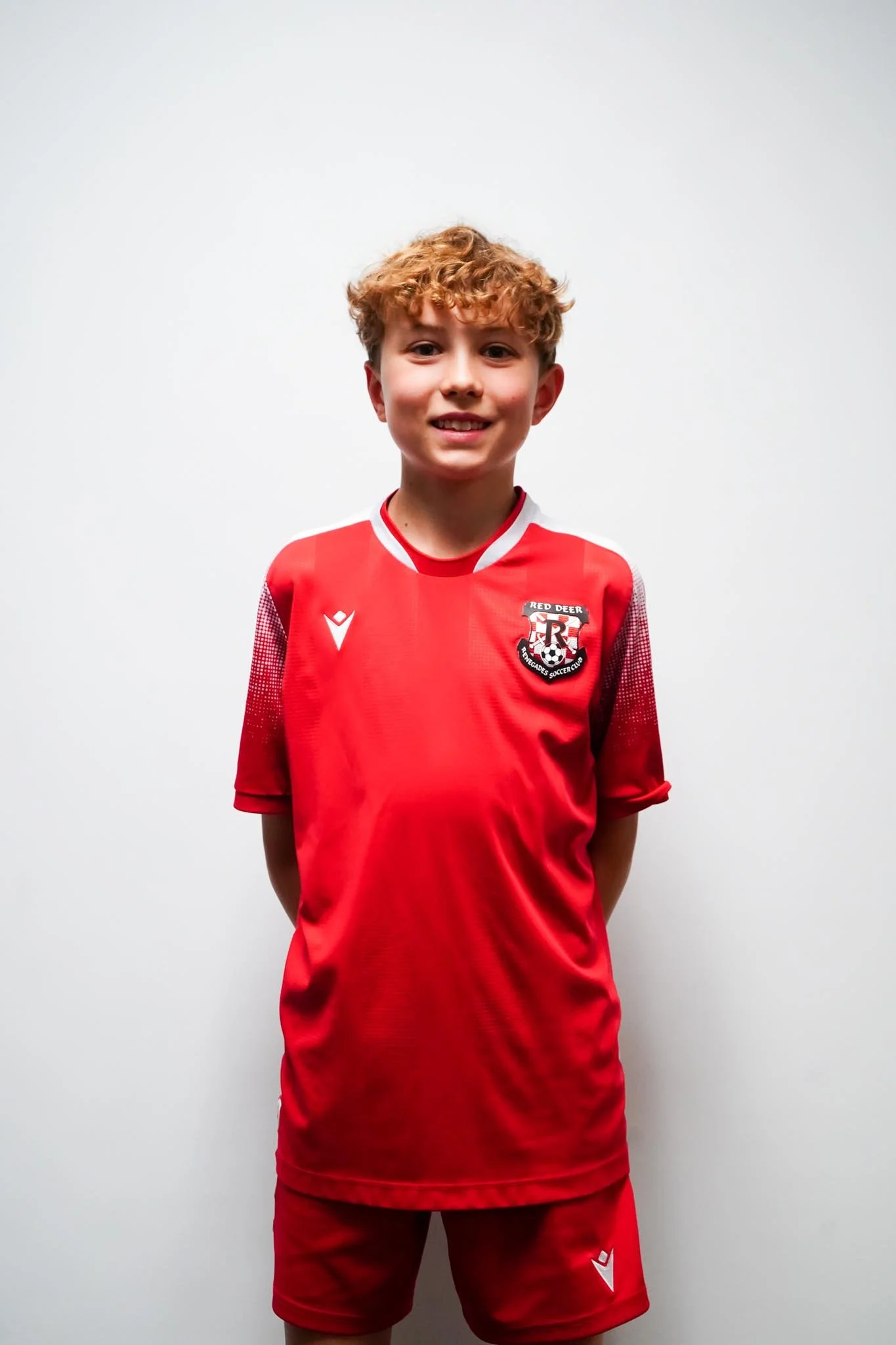 A young boy with curly red hair wearing a red soccer jersey and matching shorts, standing against a plain white background.