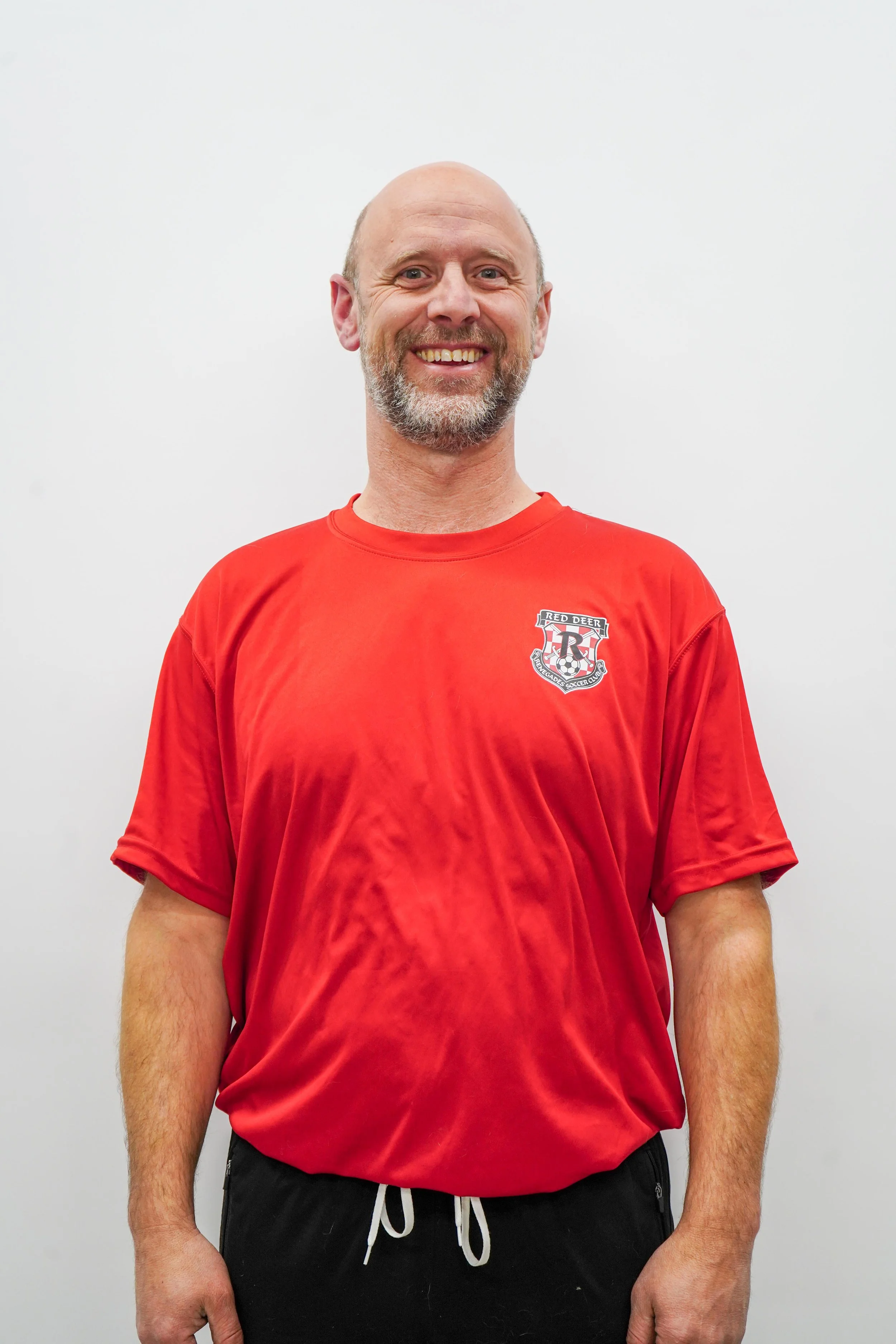 A smiling man with a beard and shaved head wearing a red sports t-shirt with a logo on the chest, standing against a plain white background.