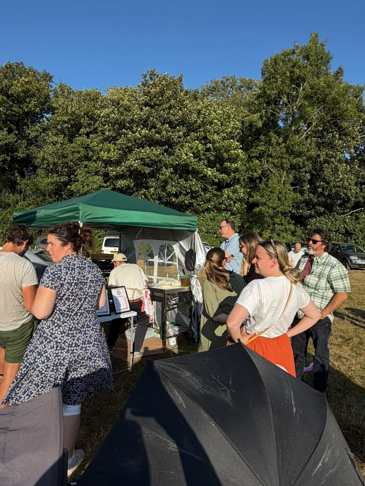 Group of people gathered outdoors at a market or event under a green canopy tent, with trees and parked cars in the background, sunny weather with a clear blue sky.