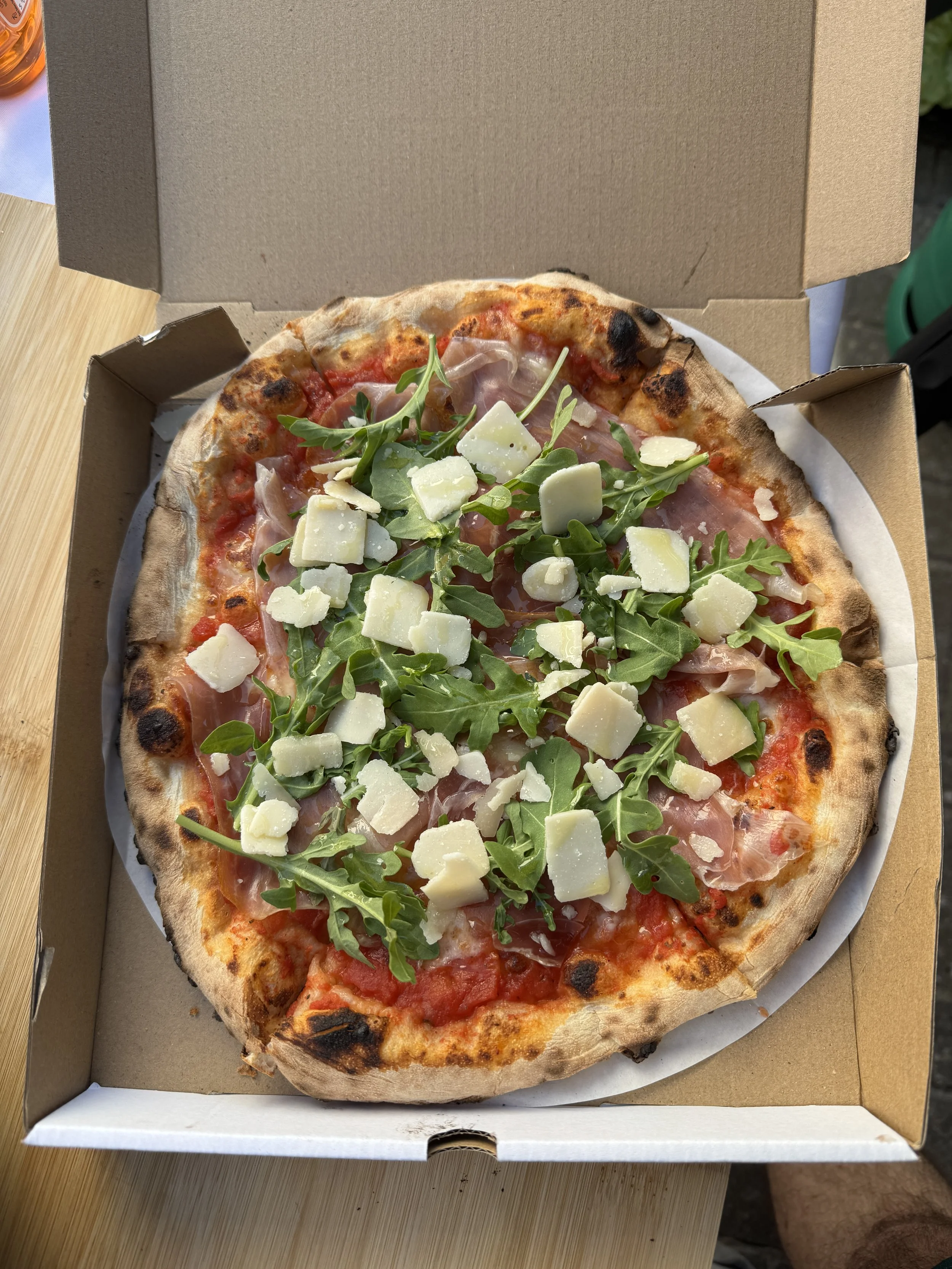 A pizza in a cardboard box with arugula, cheese, and thin slices of prosciutto, on a wooden table.