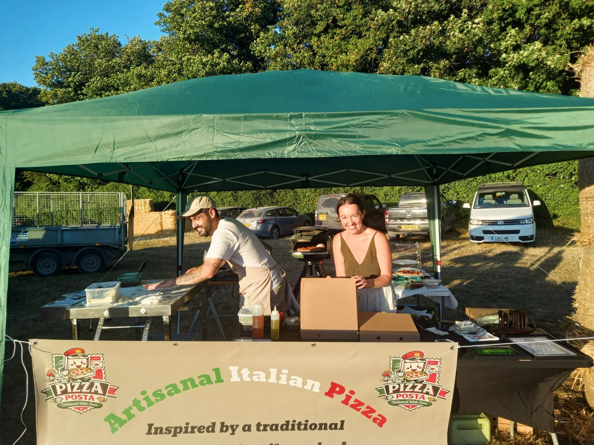Two people, a man and a woman, are standing behind a pizza stand at an outdoor event. The stand has a banner advertising artisanal Italian pizza inspired by traditional recipes. The man is wearing a baseball cap and appears to be preparing food, while the woman is smiling at the camera with a food grill in front of her. There are tents, parked cars, and trees in the background.