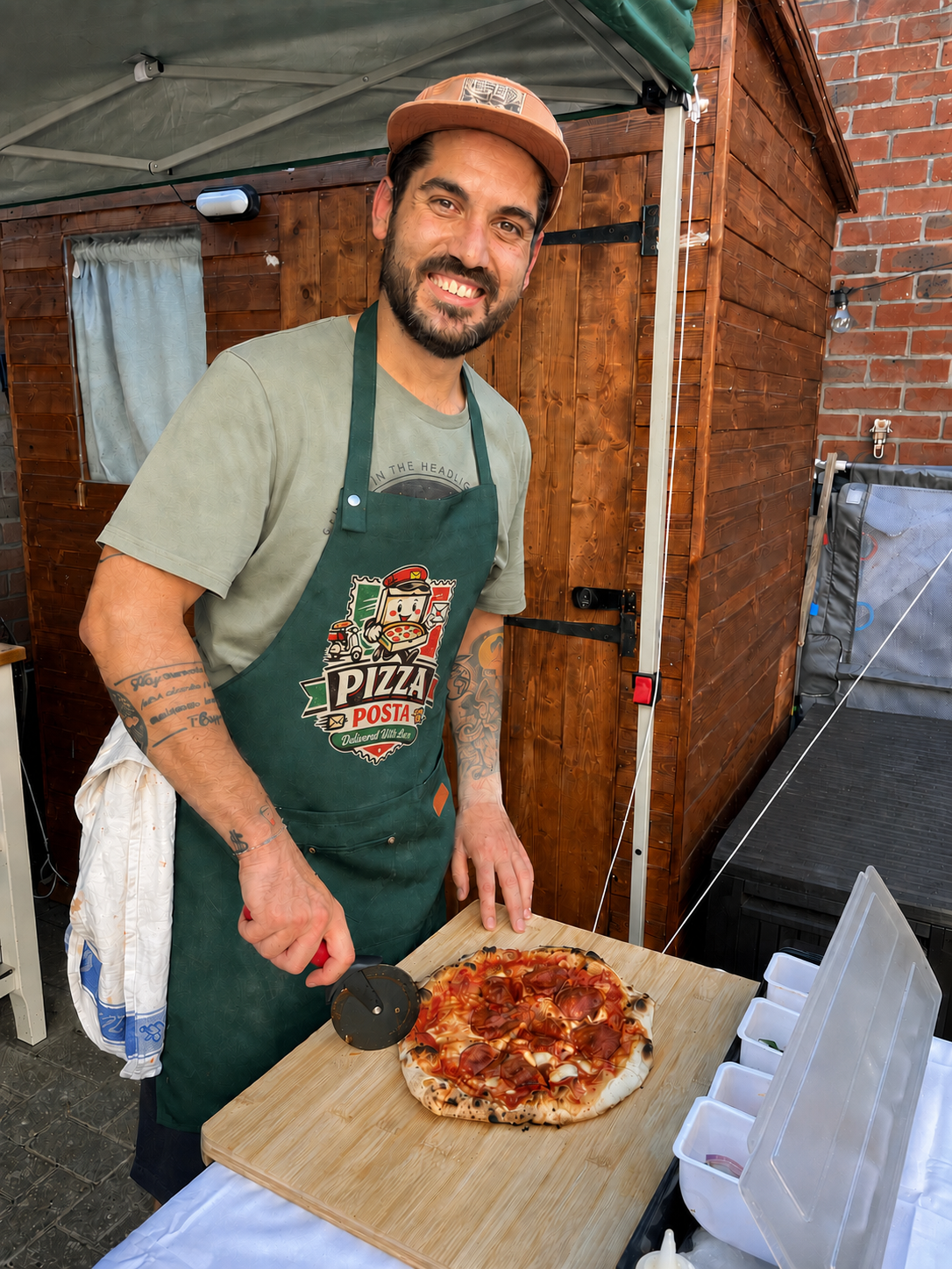 A smiling man is slicing a freshly baked pizza on a wooden cutting board. He is wearing a green apron with a pizza logo, a beige t-shirt, and a light brown cap. The background features a wooden shed and brick wall, indicating an outdoor setting.