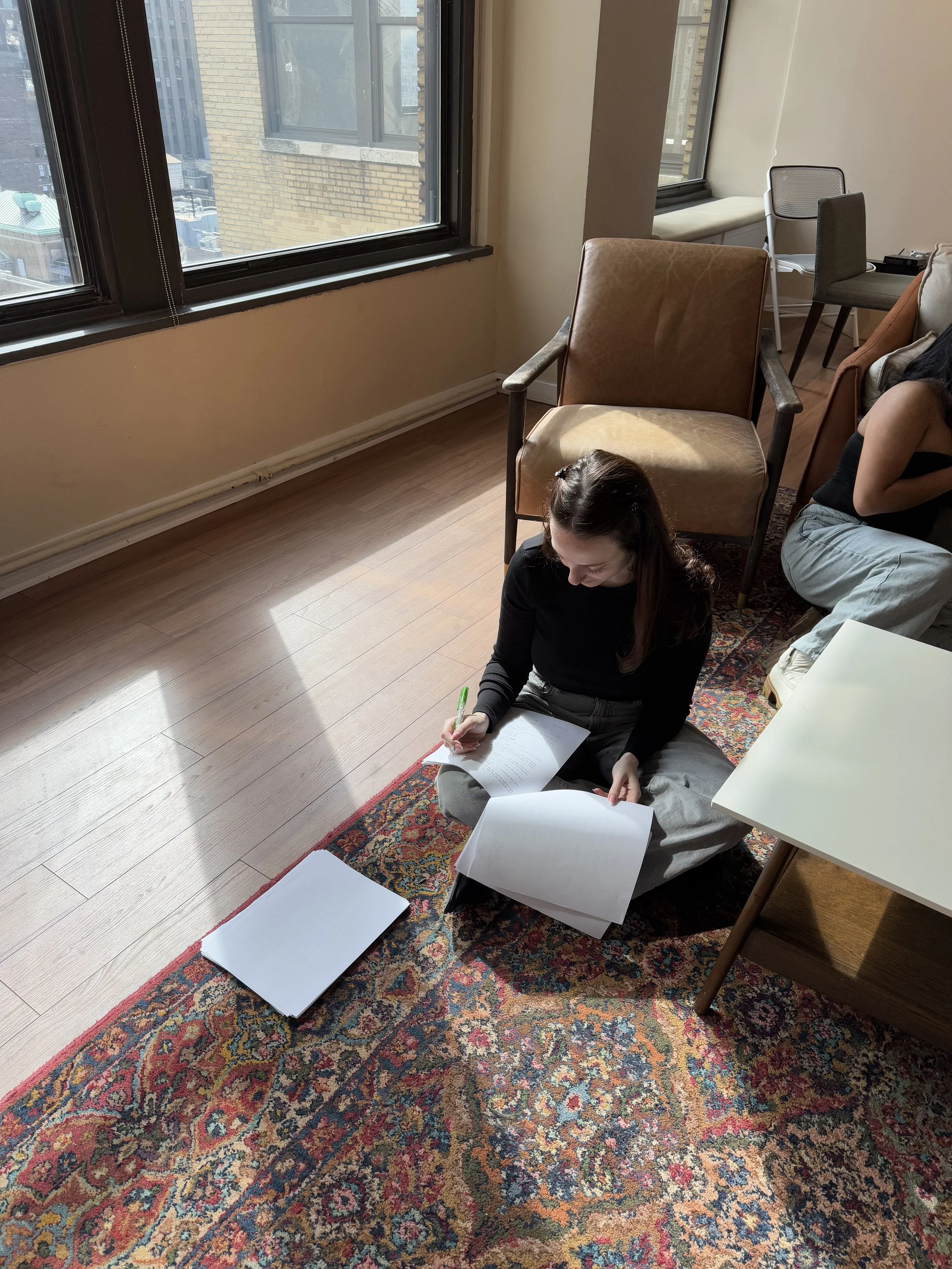 A young woman with dark hair and gray pants sitting cross-legged on an ornate rug, writing in a notebook with a green pen. She is next to a white table and is surrounded by chairs and other people in a room with large windows showing a cityscape with