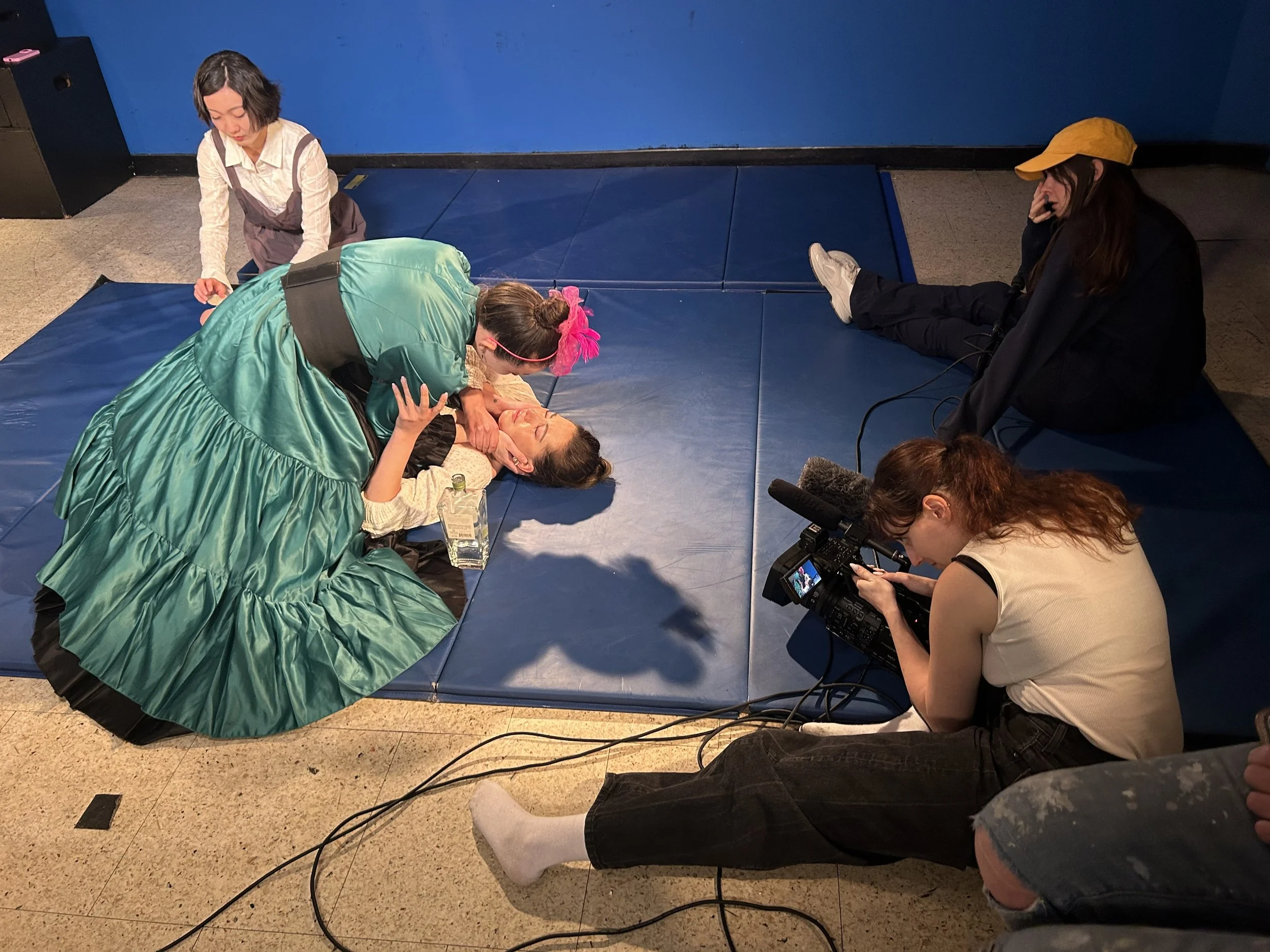 People filming a scene on a padded blue mat, with a woman lying down and another woman in a green dress leaning over her, while a girl with pink hair and headband assists. An actress sits nearby, and a crew member with curly hair and a white shirt fi