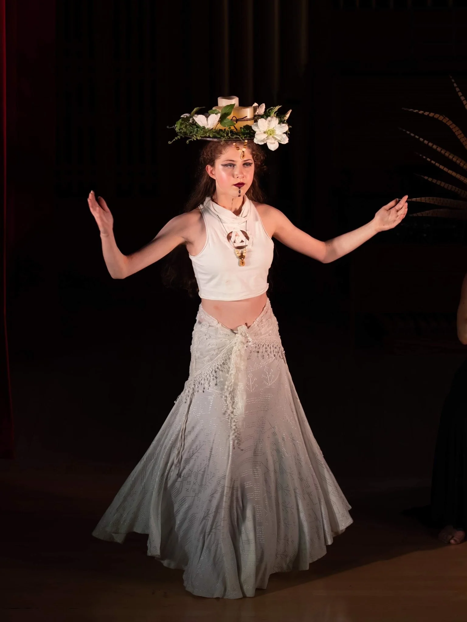 A woman dressed in a bohemian style with a wide, decorated hat, white sleeveless top, and long flowing skirt dancing on a stage.