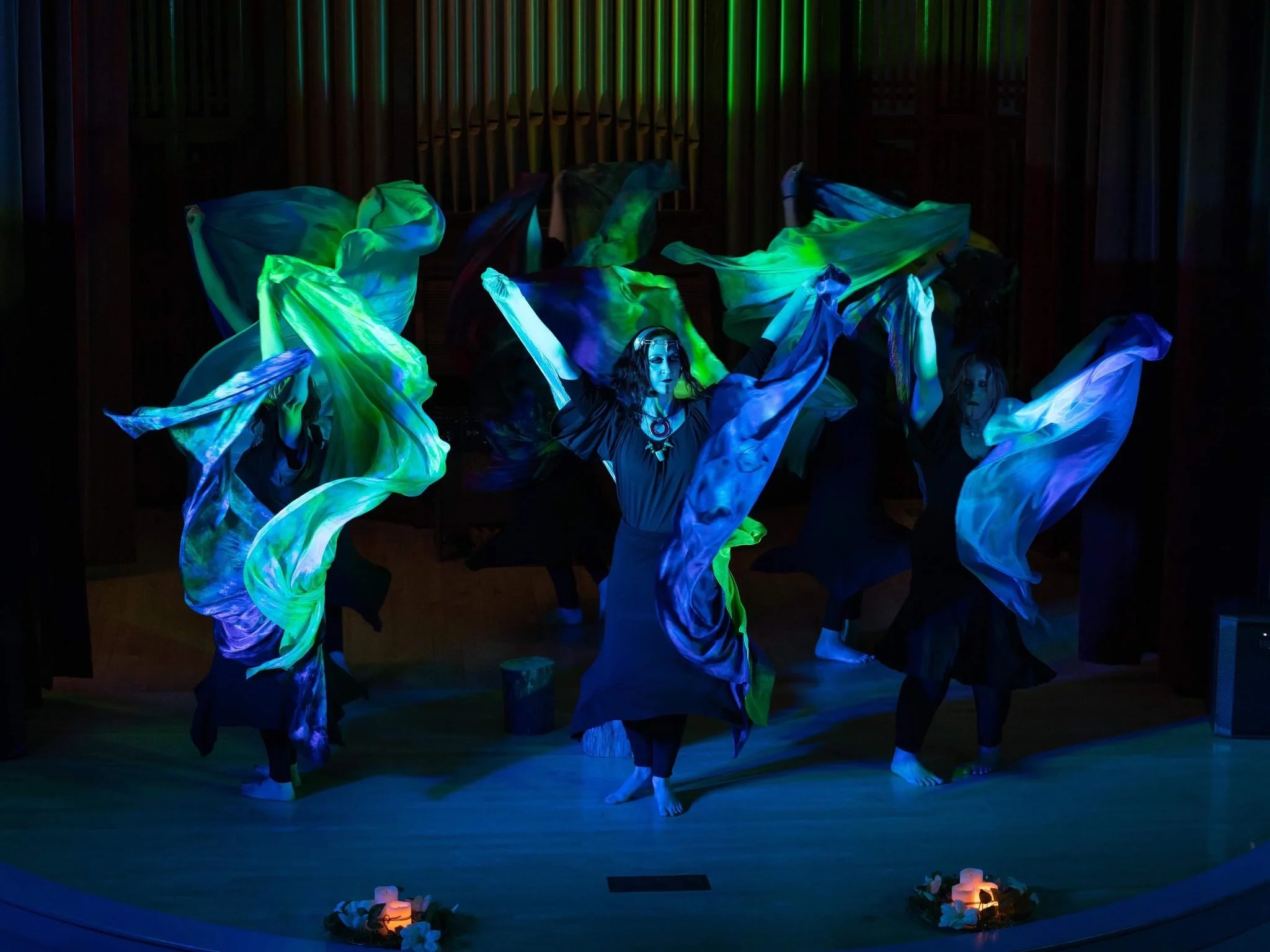 Performers dancing with flowing blue and green fabric on stage, illuminated by colorful lights.