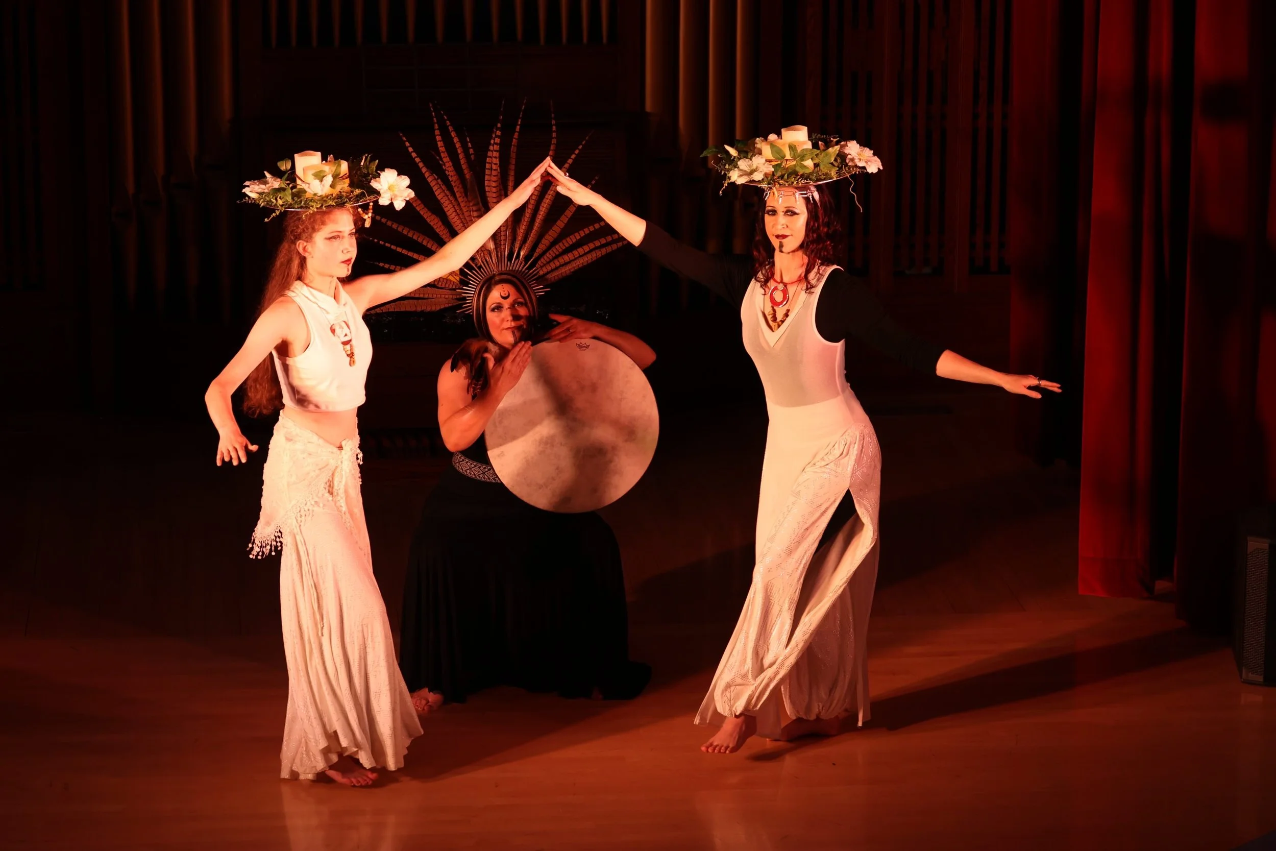 Three women performing a dance on stage, with the two women in front holding hands and the woman in the middle playing a large gong, all wearing elaborate costumes and headpieces.