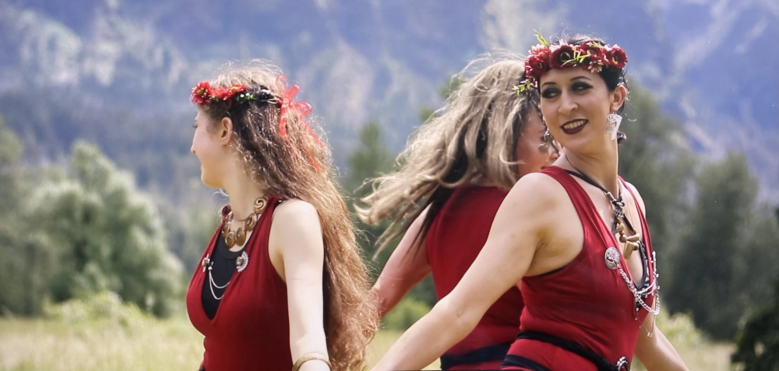 Three women wearing red dresses and floral headbands, dancing outdoors with mountains and trees in the background.
