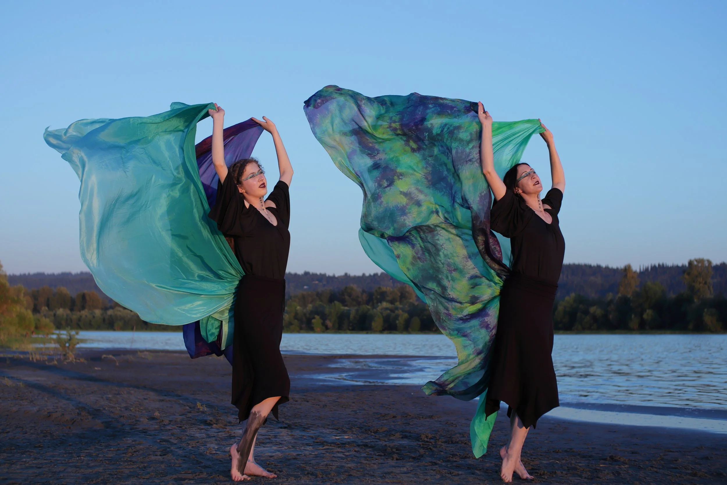 Two women in black dresses holding large, colorful fabric pieces, standing barefoot on a beach by the water with a forest in the background during sunset.