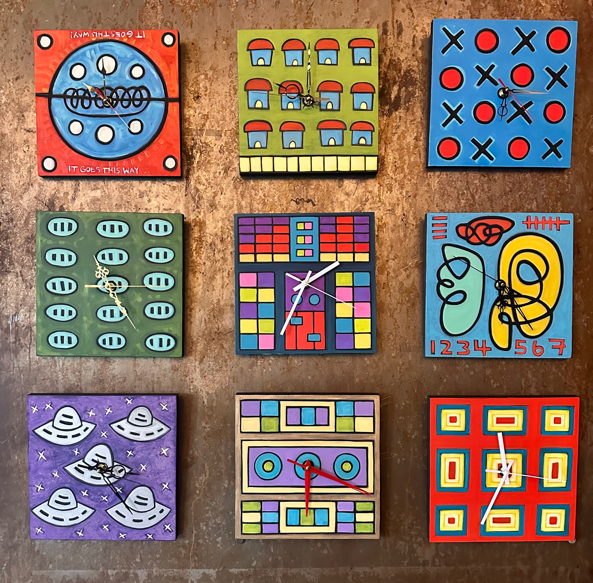 photography of nine brightly painted clocks with bold lines, colors, and various themes and shapes against a copper gallery wall on Capitol Hill Seattle. The center clock handpainted clock looks like a beat production machines used to make samples