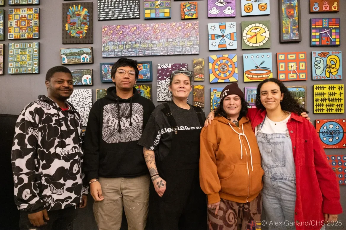 Photography of four intergenerational artists of color of Black, Indigenous, Asian decent standing and smiling in front of a wall at an art gallery on Seattle's Capitol Hill Pike Pine corridor. The wall is full of brightly painted artwork.