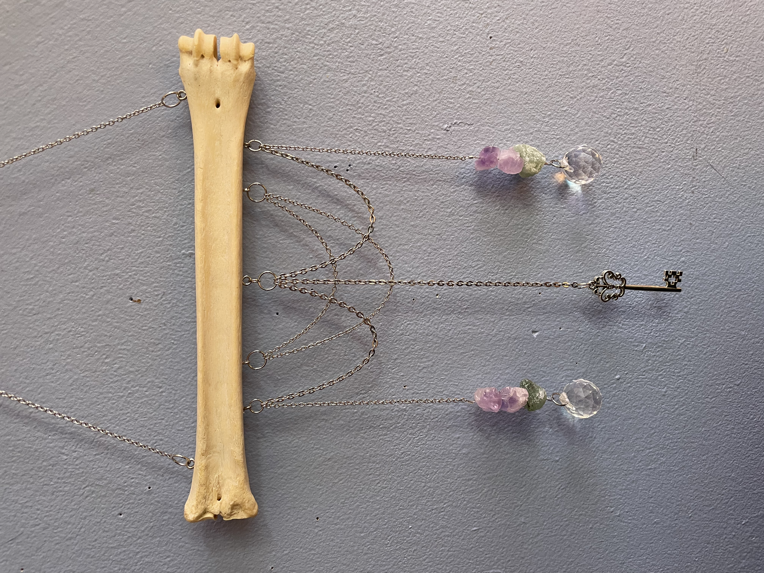 A crystal suncatcher mounted on a large foraged deer bone with decorative silver chains, rough amethyst chunks, raw green aventurine beads, an antique silver key, and two reflective prisms creating a disco ball light effect.