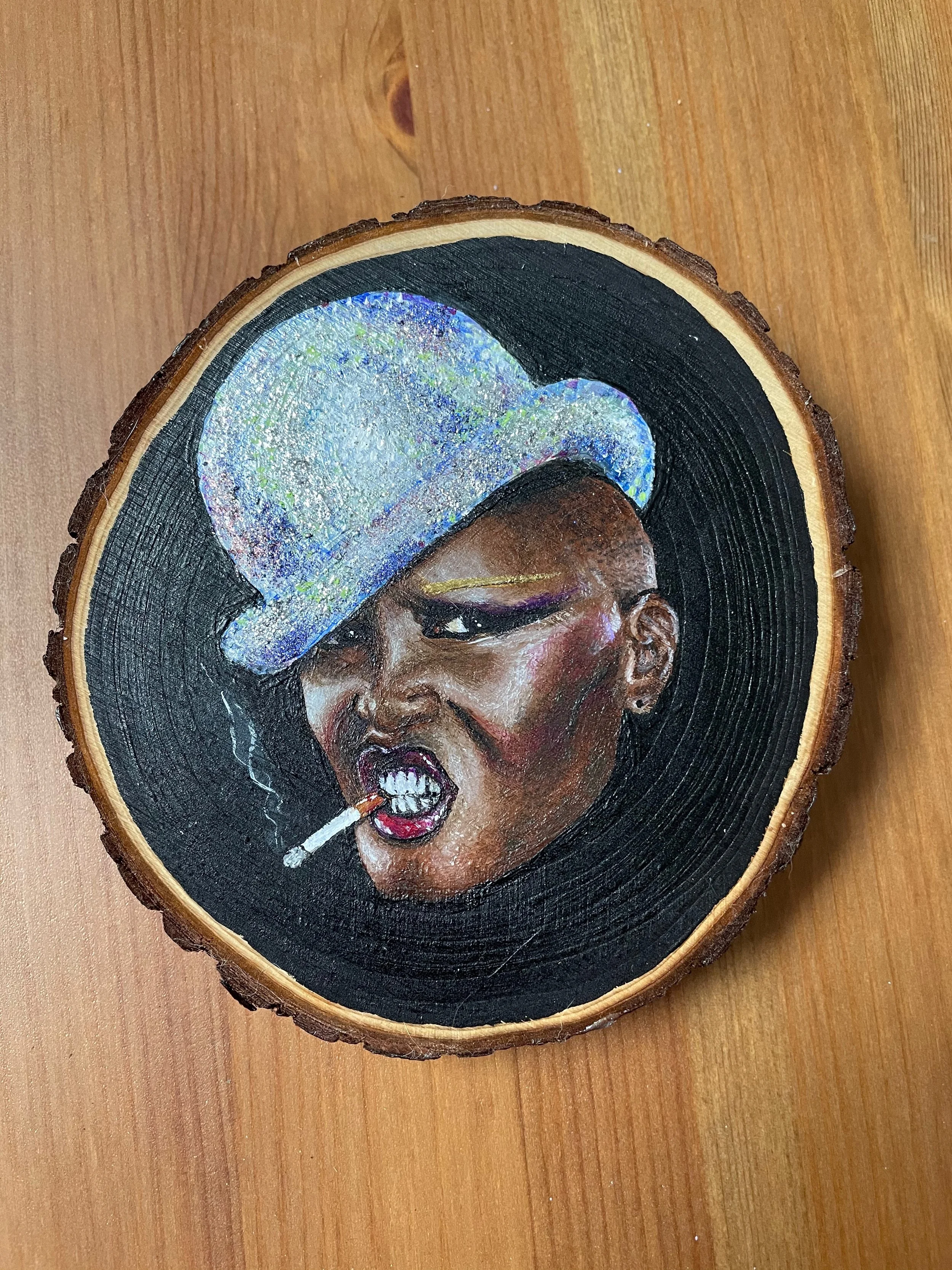A small portrait of Grace Jones painted in acrylic on a round slice of wood. The image is based on the Jean-Paul Goude photo of the artist in a dark void, with a glittering silver bowler hat and a cigarette clenched between her teeth.