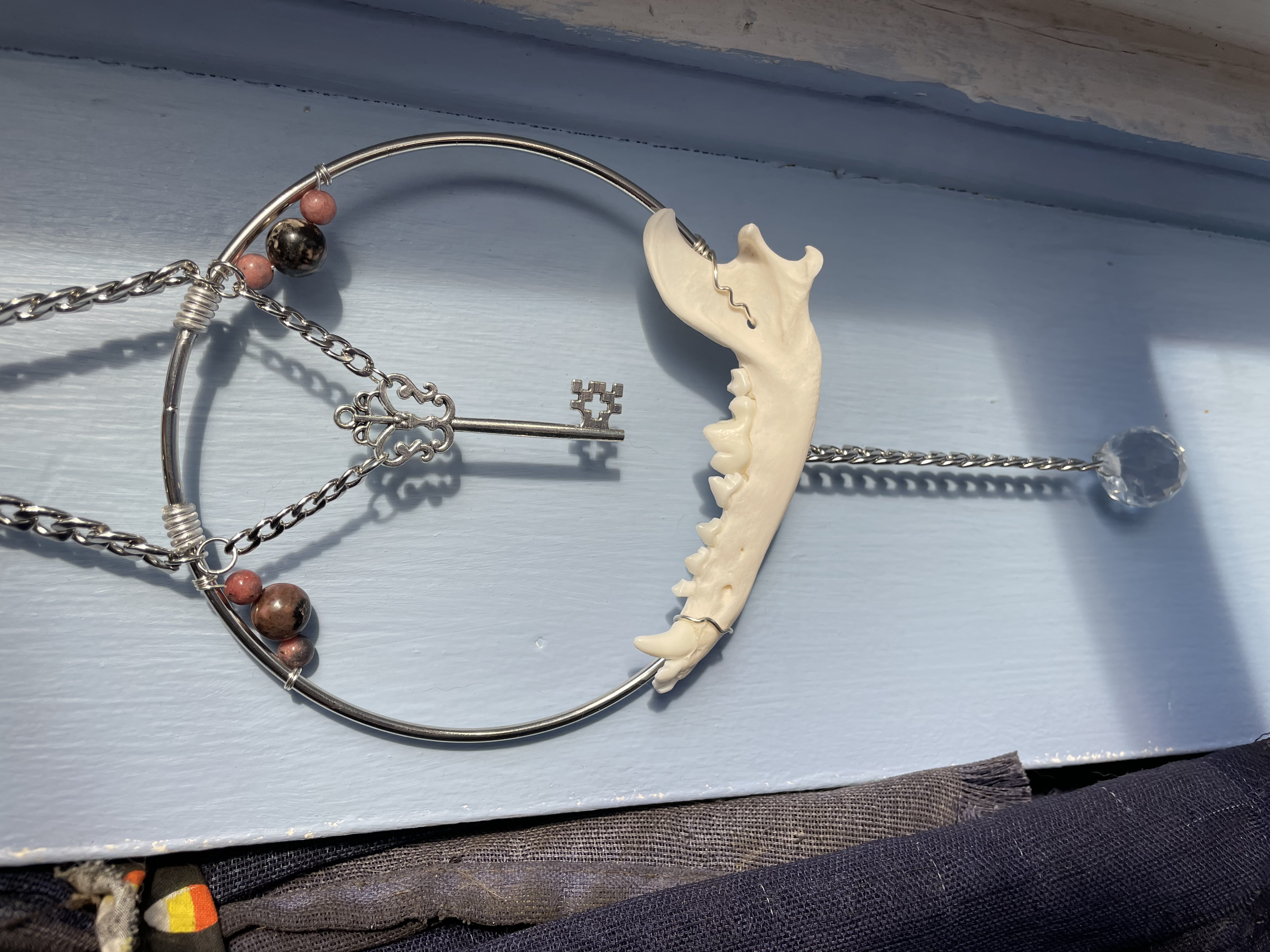 A ring-shaped crystal suncatcher featuring the jawbone of a red fox, rhodonite crystal beads, an antique silver key, chains, and reflective prisms creating a disco ball light effect.