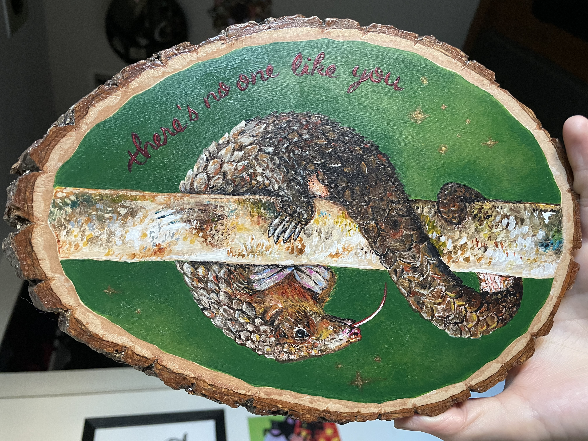 An image of a pangolin curled around a tree trunk, hand-painted in acrylic on a large oval-shaped wood slice. The creature peeks out from a forest green background with the phrase "there's no one like you" painted around in copper script.