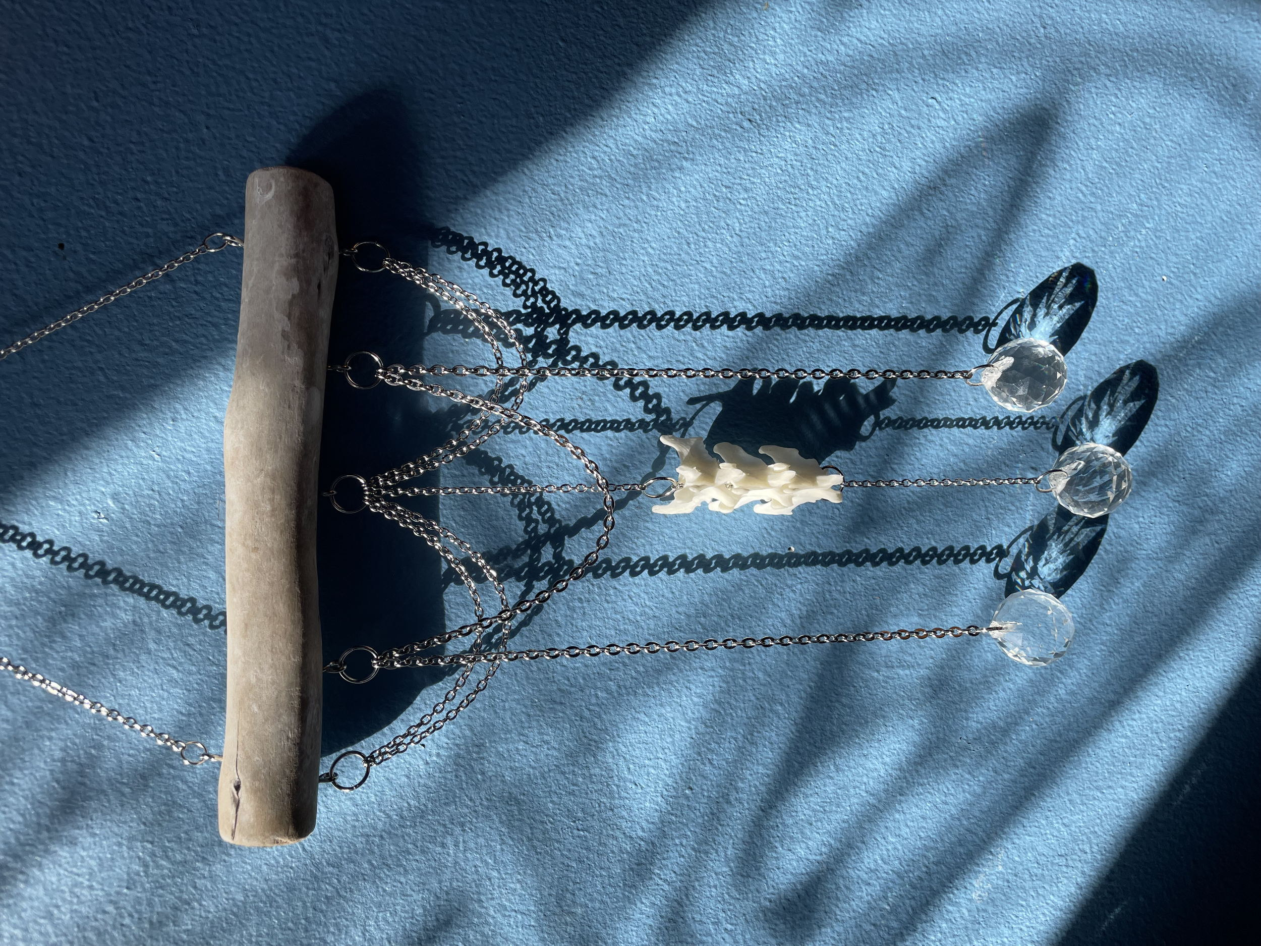 A crystal suncatcher mounted on foraged driftwood with layered decorative chains, three mink vertebrae, and three reflective prisms creating a disco ball light effect.