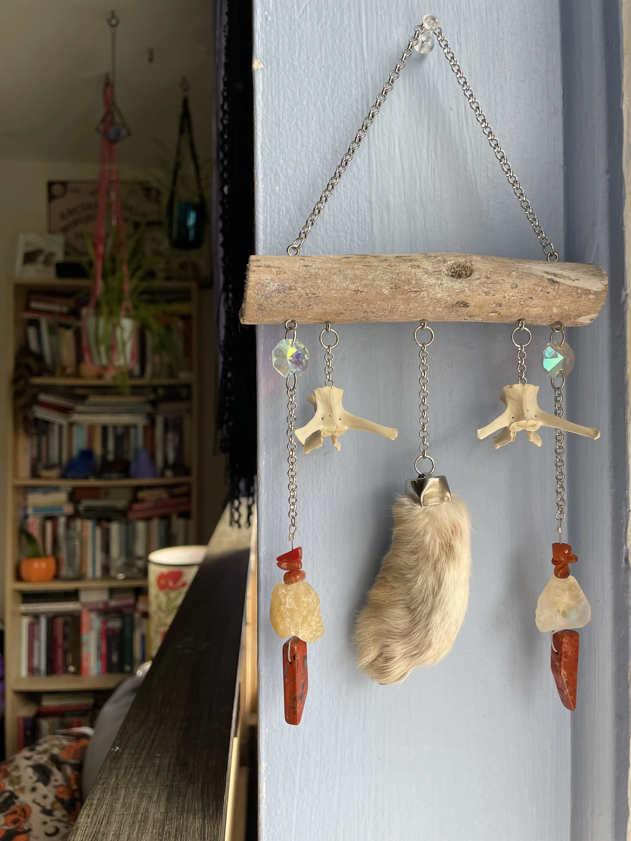 A crystal suncatcher mounted on driftwood featuring a rabbit's foot, two rabbit vertebrae, raw citrine pieces, red jasper, and red tiger's eye, plus reflective prisms creating a disco ball light effect.