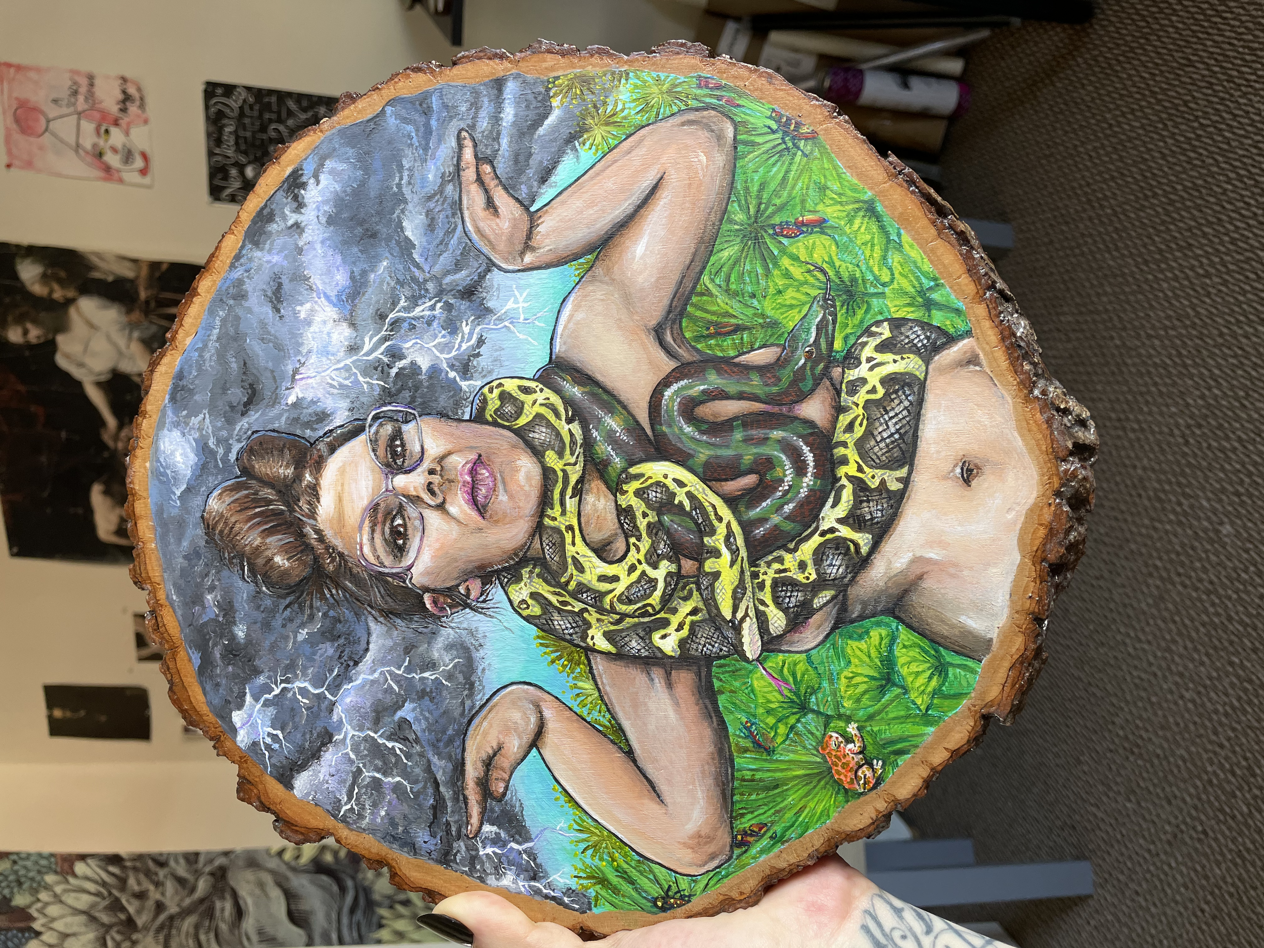A large portrait of a woman with snakes draped across her bare chest, painted in acrylic on a round wooden slab. The background features thunder clouds, bright green Egyptian plants, beetles, and frogs.