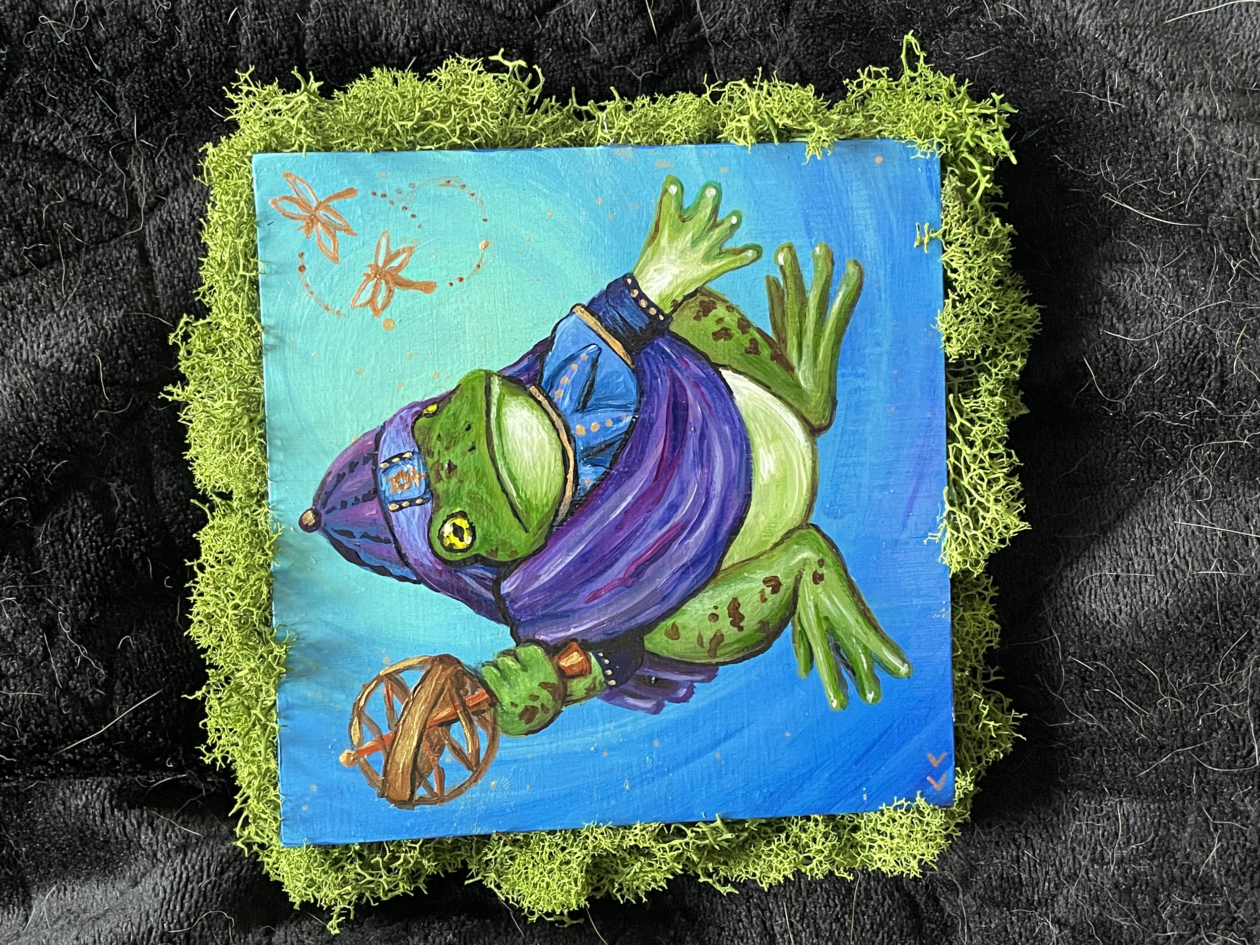 A small painting of a plump green frog presented as the thrice great polymath Hermes Trismegistus, with a purple hat and sash, holding a caduceus, set against a blue background with floating dragonflies. The painting is framed by bright green moss.