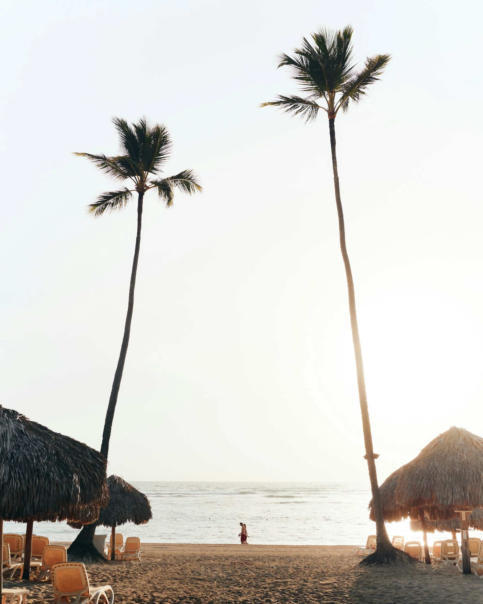 Sunset beach scene with two tall palm trees and thatched umbrellas, empty chairs, and a person walking along the shoreline.