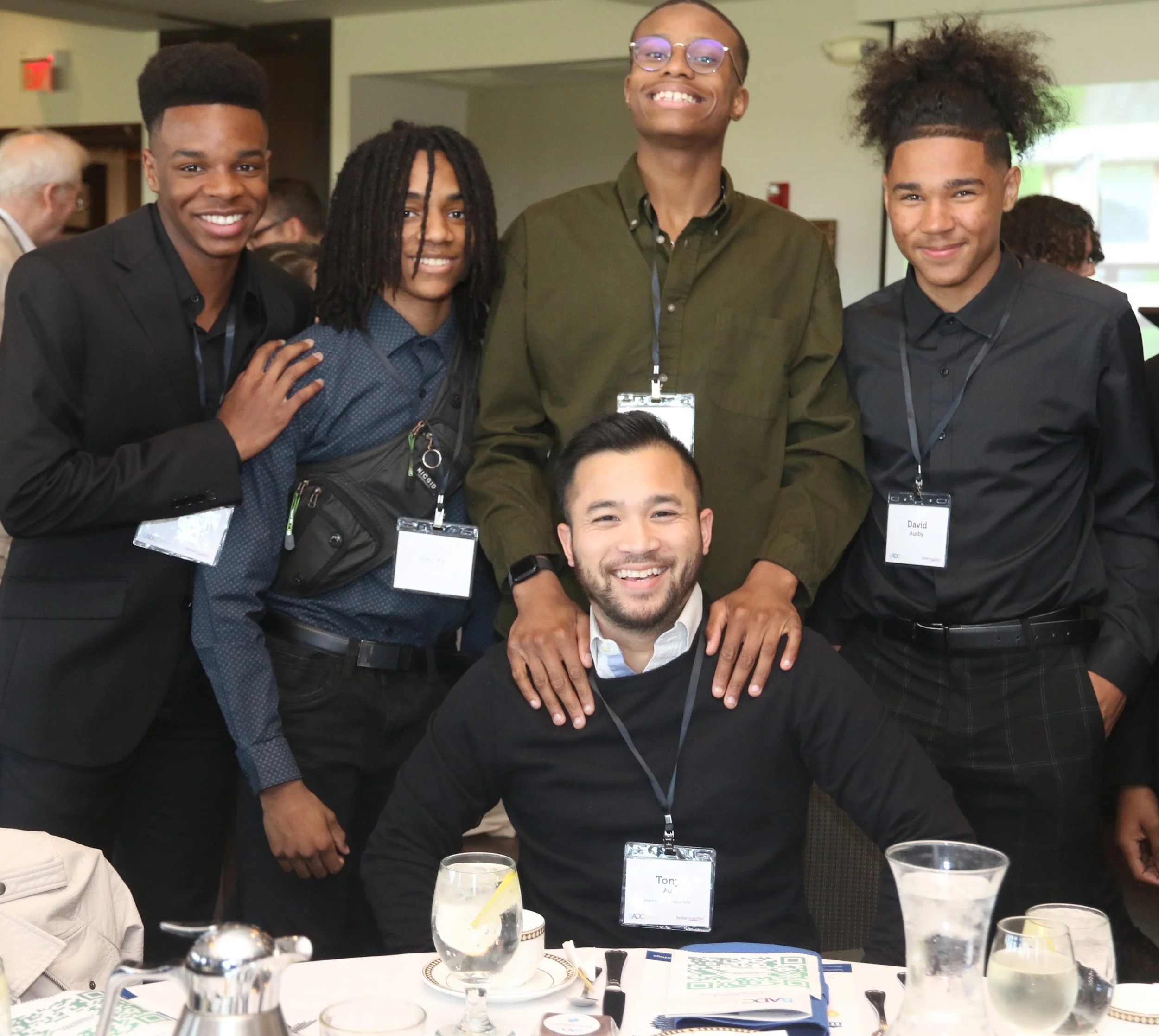 Group of five young men smiling at a party or conference, sitting and standing around a table with drinks, wearing name tags, in a well-lit room.