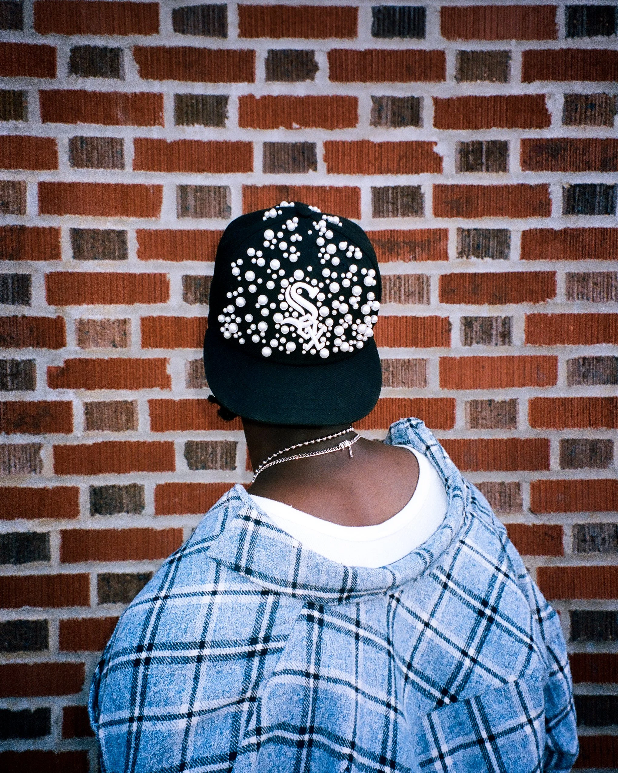 Person wearing a black baseball cap decorated with white beads, a white T-shirt, and a blue plaid shirt, standing in front of a brick wall.