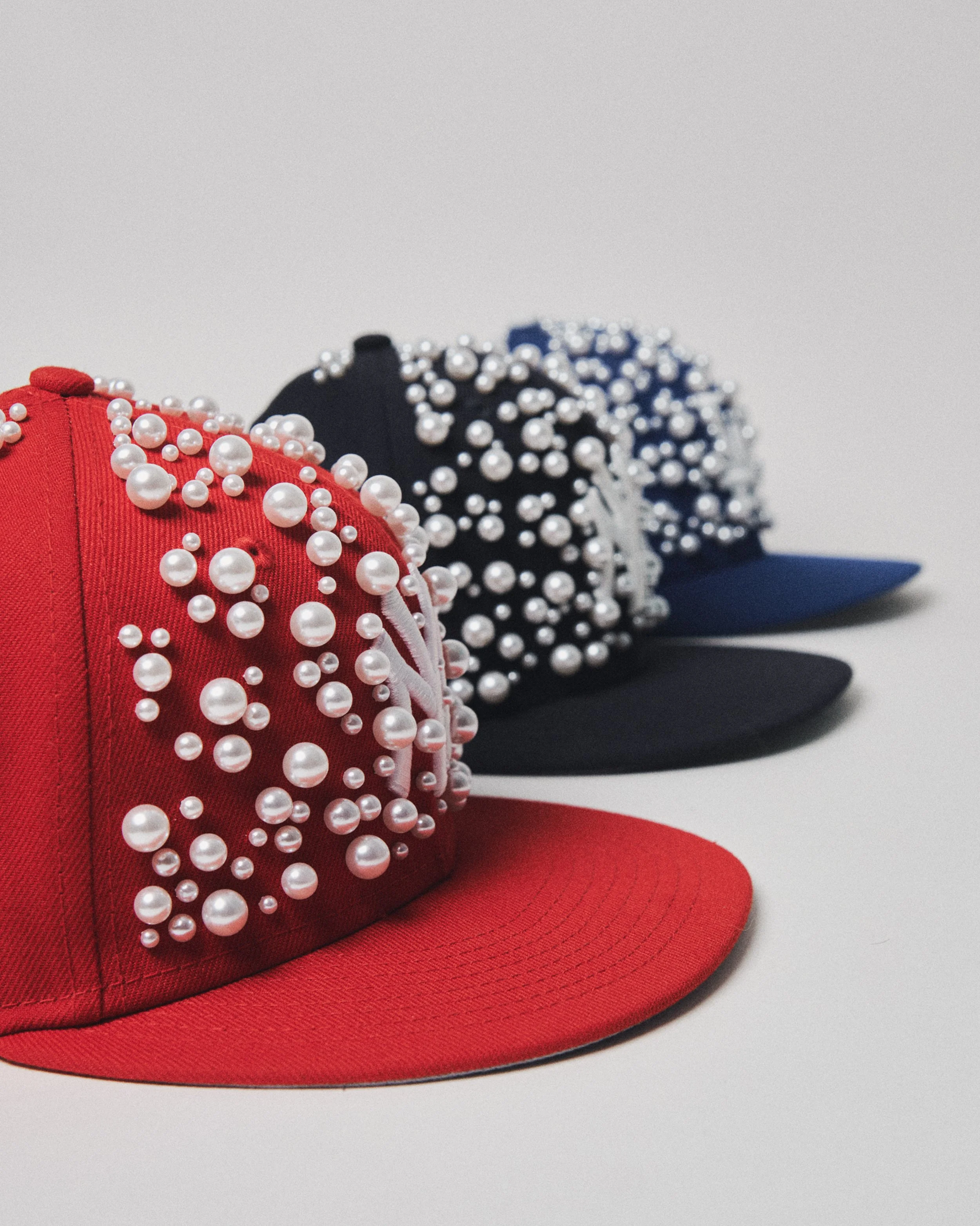 Three baseball caps decorated with white pearl beads, in red, black, and blue, arranged in a line on a white surface.
