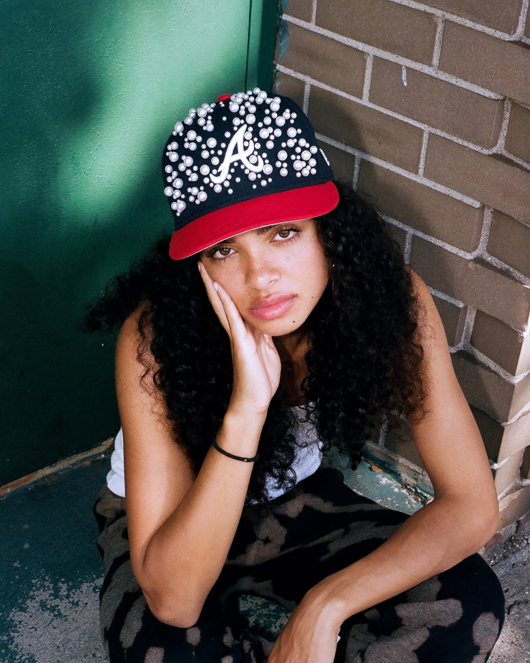 Young woman with curly black hair wearing a black baseball cap decorated with white pearls and a red brim, sitting against a brick wall and green door.
